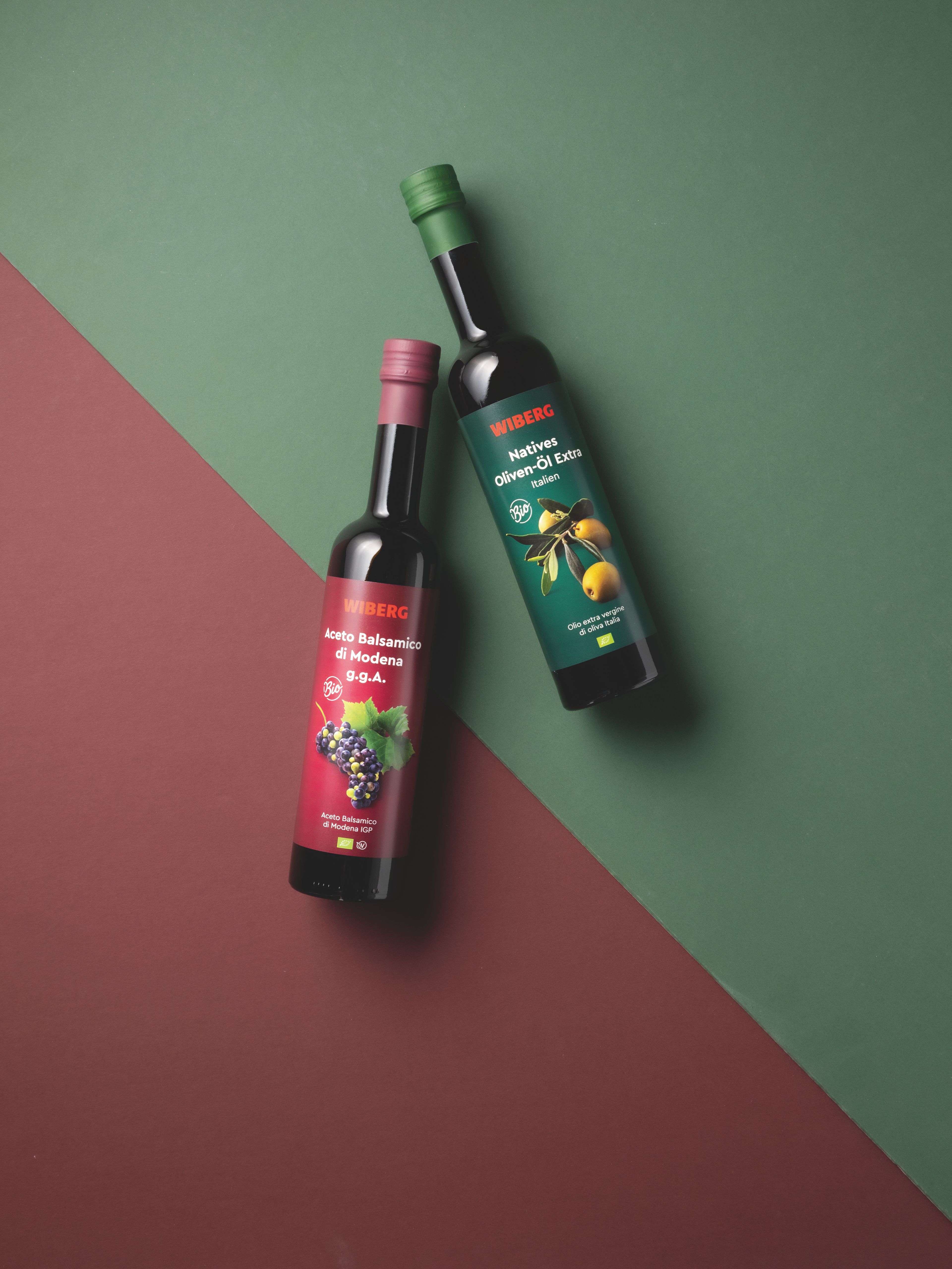WIBERG BIO Aceto Balsamico di Modena g.g.A. in dunkler 500 ml Flasche mit dunkelrotem Etikett und WIBERG BIO Natives Oliven Öl Extra Italien in dunkler 500 ml Flasche mit dunkelgrünem Etikett auf diagonal geteiltem Hintergrund in Dunkelrot und Dunkelgrün.