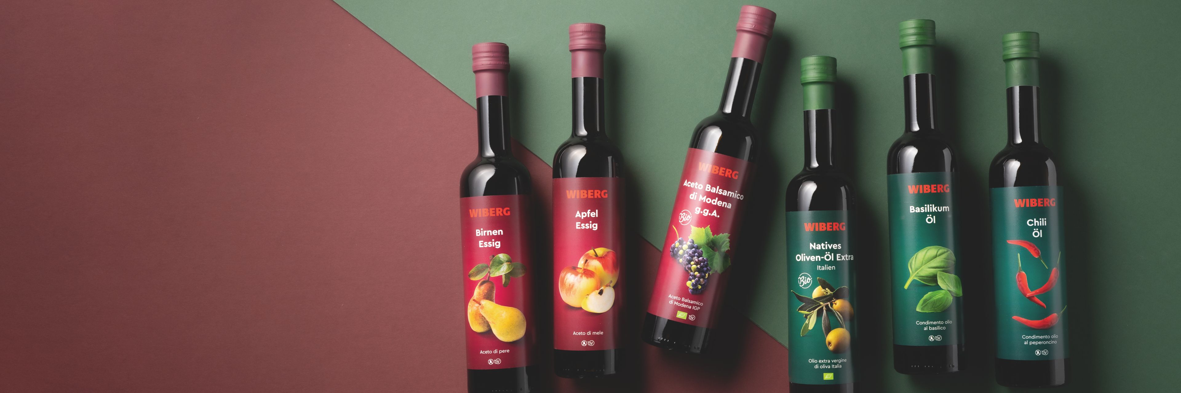 WIBERG Birnen‑Essig, WIBERG Apfel‑Essig und WIBERG Aceto Balsamico di Modena g.g.A. mit dunkelroten Etiketten sowie WIBERG Natives Oliven‑Öl Extra Andalusien, WIBERG Basilikum‑Öl und WIBERG Chili‑Öl mit dunkelgrünen Etiketten in dunklen 500‑ml‑Flaschen auf diagonal geteiltem Hintergrund in Dunkelrot und Dunkelgrün.