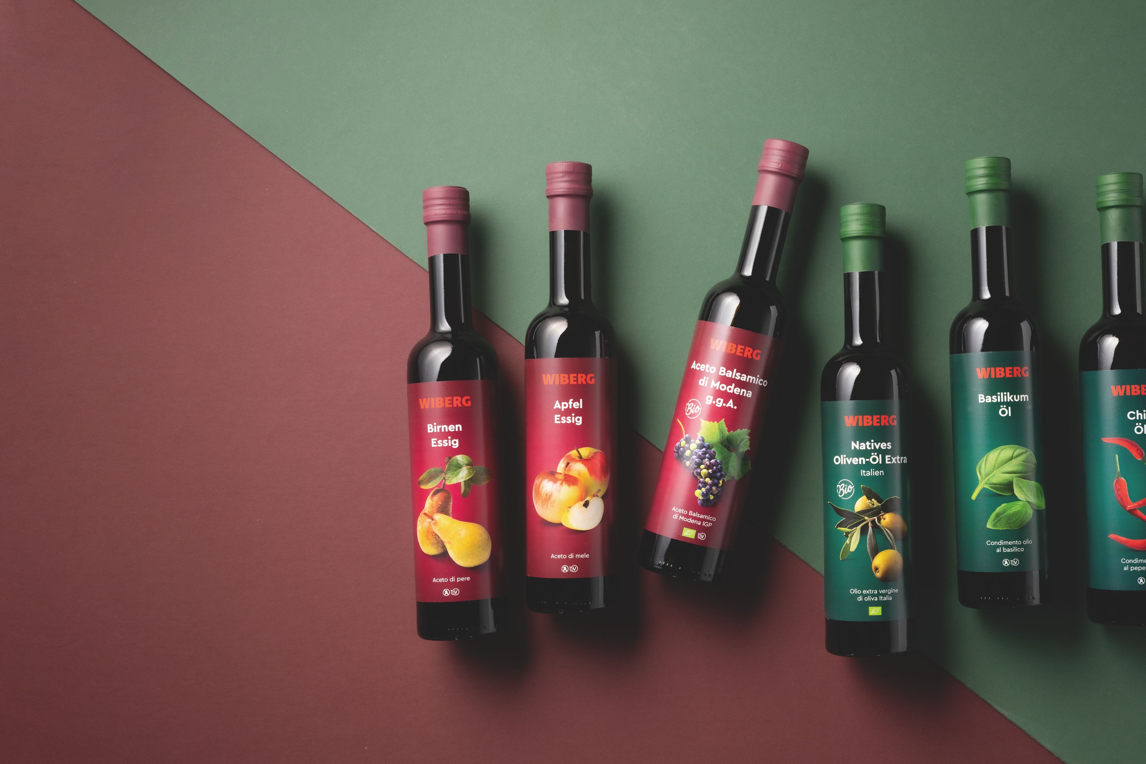 WIBERG Birnen‑Essig, WIBERG Apfel‑Essig und WIBERG Aceto Balsamico di Modena g.g.A. mit dunkelroten Etiketten sowie WIBERG Natives Oliven‑Öl Extra Andalusien, WIBERG Basilikum‑Öl und WIBERG Chili‑Öl mit dunkelgrünen Etiketten in dunklen 500‑ml‑Flaschen auf diagonal geteiltem Hintergrund in Dunkelrot und Dunkelgrün.