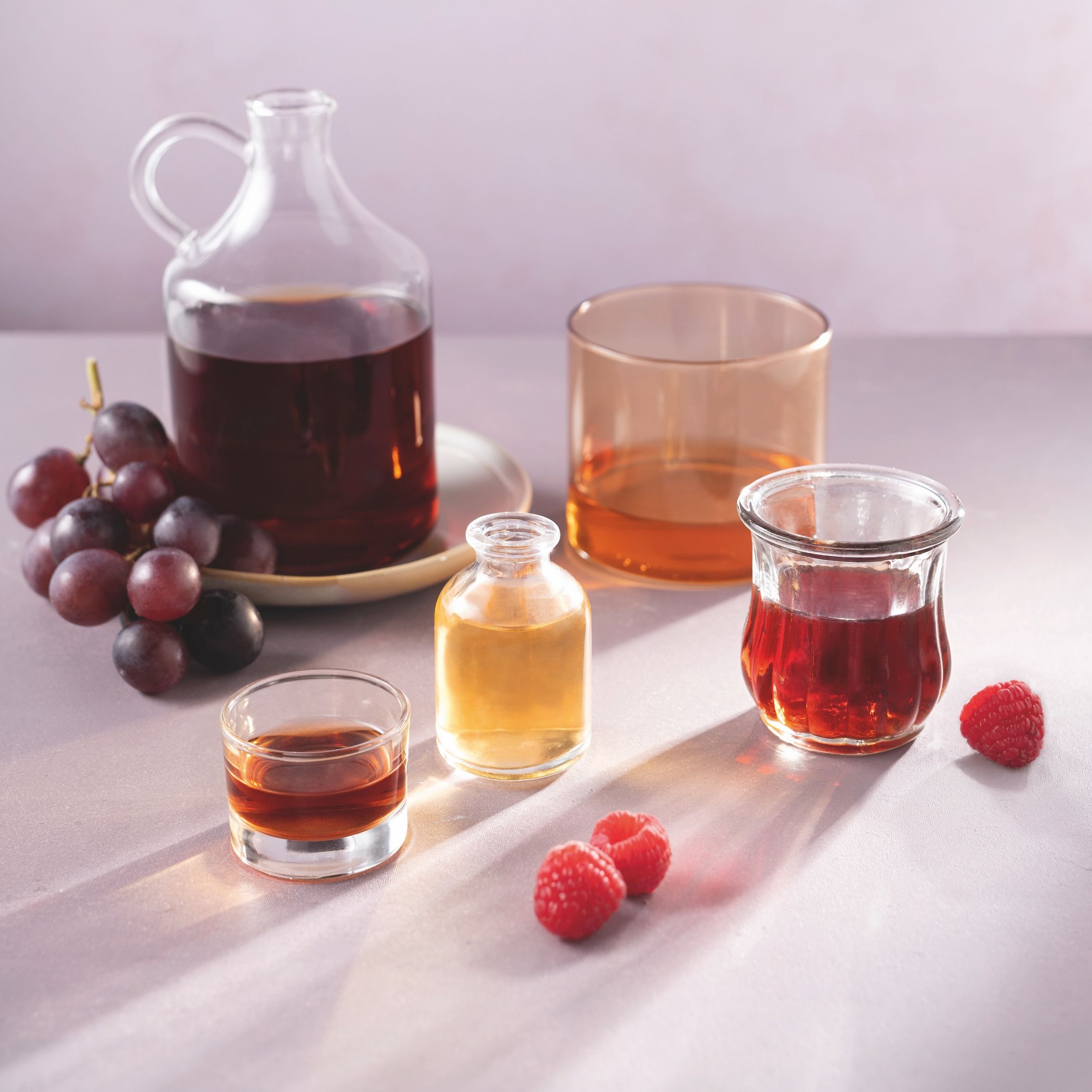 Verschiedene Gefäße mit WIBERG Premium Essigen vor hellem grau lila Hintergrund: links eine Glaskaraffe mit Aceto Balsamico auf hellgrauem Teller mit dunkellila Weintraubenrispe, daneben ein helloranges Glas mit hellem Essig, im Vordergrund ein kleines Glas mit Essig, mittig ein Fläschchen mit hellem Apfel Essig und rechts ein orientalisches Glas mit rotem Himbeer Essig; drei Himbeeren liegen davor, Licht von hinten erzeugt Schatten und leuchtende Reflexionen durch die gefüllten Gläser.
