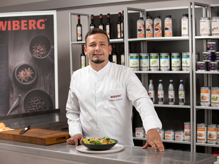 Philipp Kohlweg vom WIBERG Team Inspiration in weißer Kochjacke vor einem Regal mit WIBERG Produkten; vor ihm steht ein dekorativ angerichtetes Wok‑Gericht auf der Arbeitsfläche.