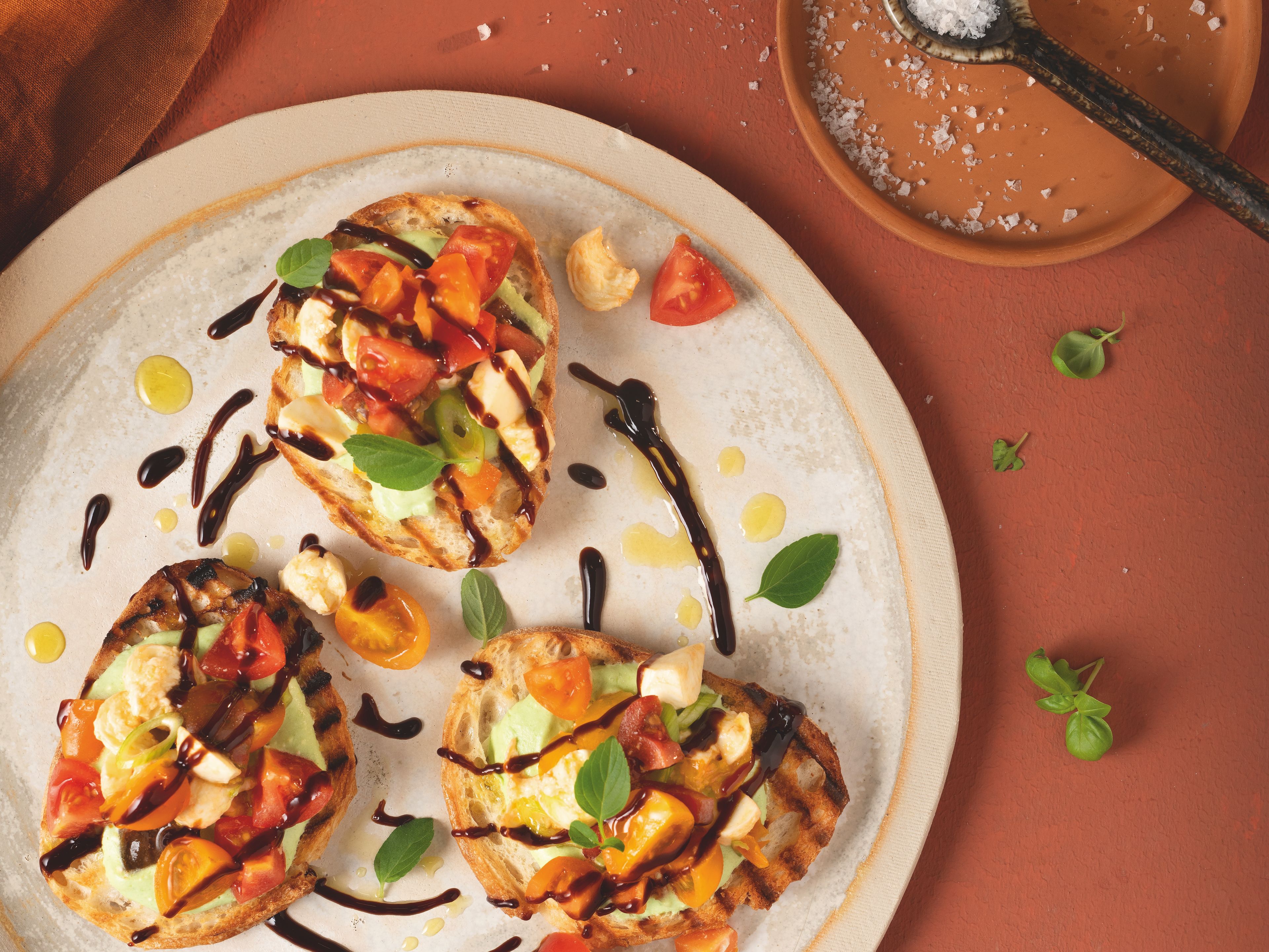 Von oben fotografierter Teller mit drei Bruschetta‑Brötchen, belegt mit rot‑ und dunkelgelben Tomaten, verfeinert mit WIBERG Würzsauce Tasty Umami und frischem Basilikum, auf rostrotem Hintergrund.