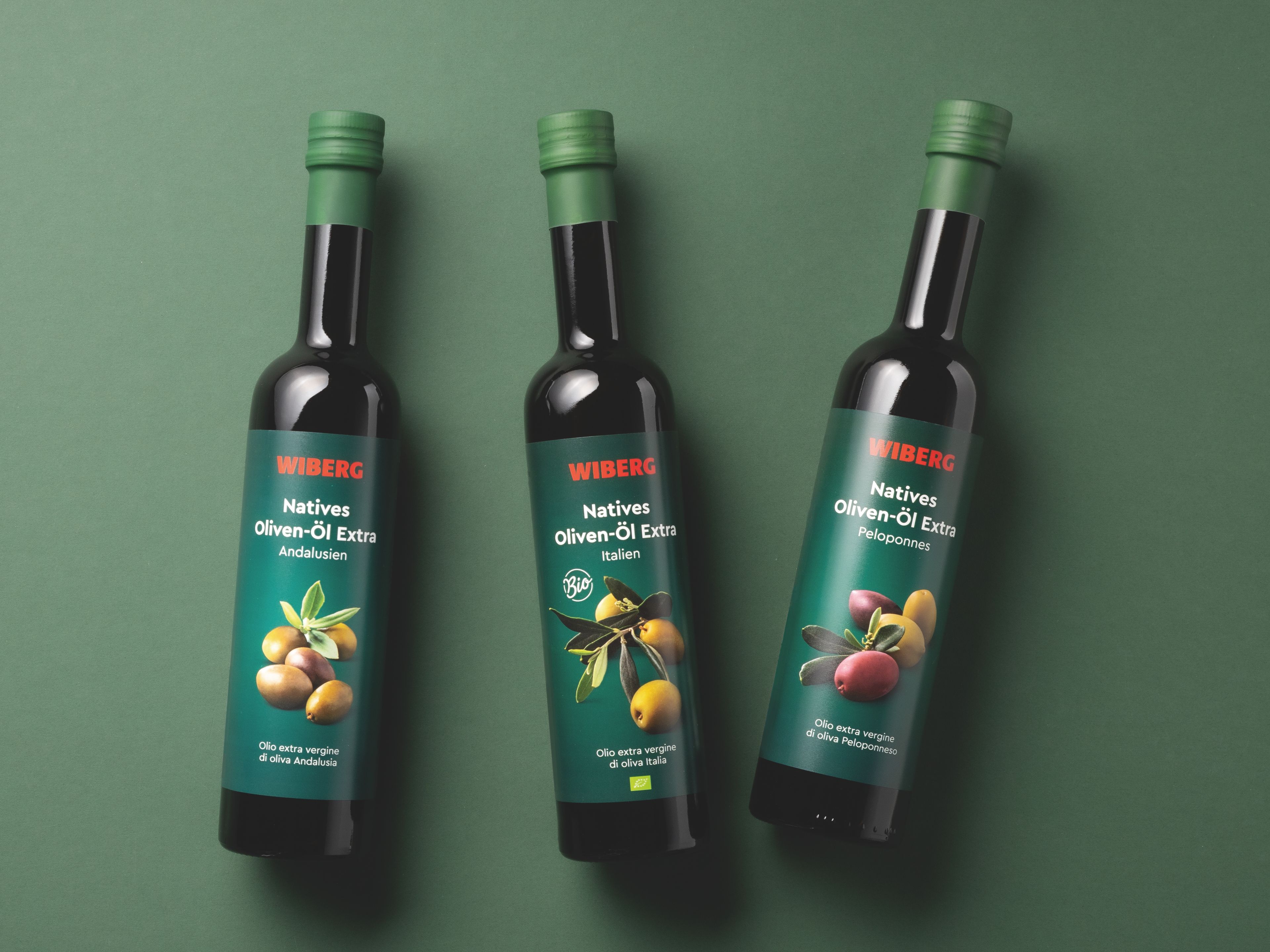 Drei dunkle 500‑ml‑Flaschen der WIBERG Premium‑Olivenöle – Natives Oliven‑Öl Extra Andalusien, BIO Natives Oliven‑Öl Extra Italien und Natives Oliven‑Öl Extra Peloponnes – nebeneinander auf dunkelgrünem Hintergrund mit neuem Etikettendesign.
