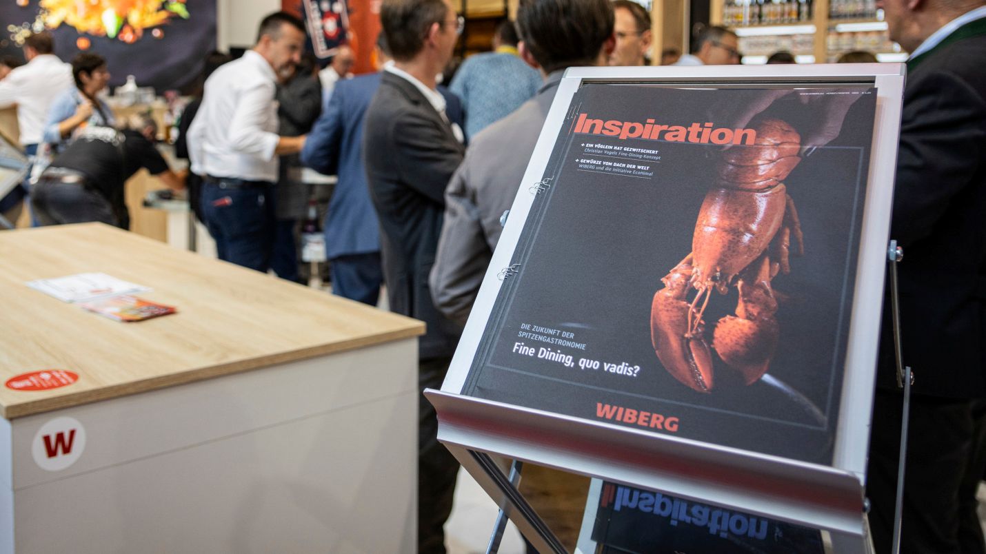 Im Querformat aufgenommenes Bild vom WIBERG Kundenmagazin „Inspiration“ mit schwarzem Titelblatt und Hummermotiv als Fine Dining Ausgabe. Das Magazin liegt auf einem Zeitschriftenständer am WIBERG Messestand einer Gastronomiemesse, im Hintergrund unscharf Besucher auf dem gut frequentierten Stand.