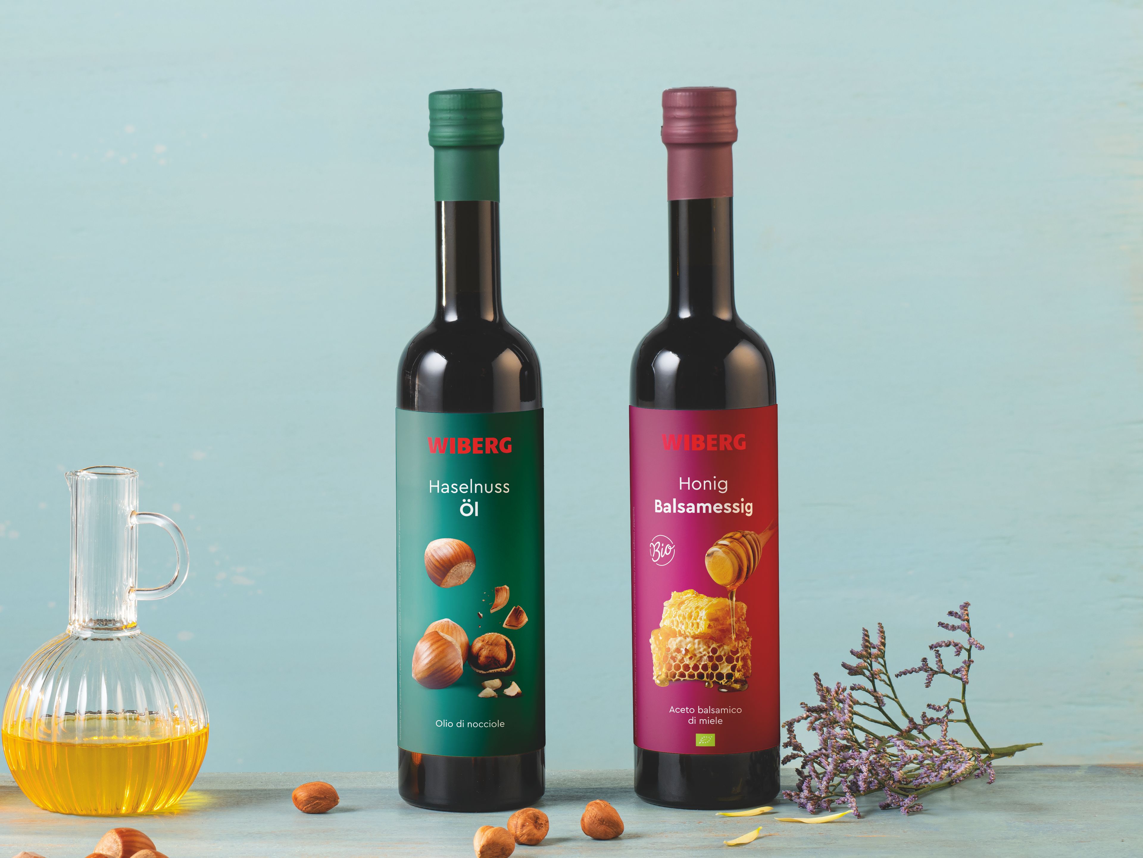 Von vorne fotografierte WIBERG Produkte auf pastellblauem Hintergrund: links Honigwabe und Haselnüsse, dahinter Karaffen mit WIBERG BIO Honig Balsamessig und Haselnuss Öl; rechts zwei 500 ml Flaschen mit grünem Haselnuss Öl Etikett und rotem BIO Honig Balsamessig Etikett.