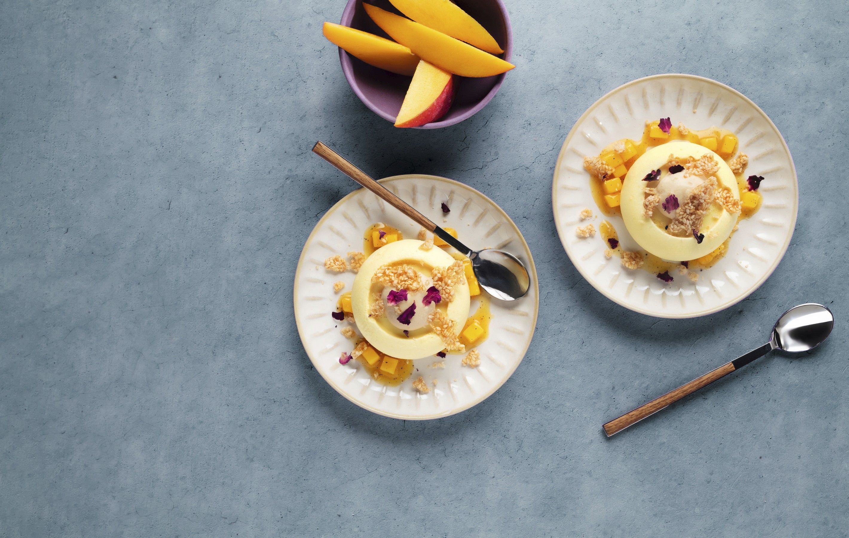 Zwei kleine helle Dessertteller mit rundem Frozen‑Mangojoghurt, angerichtet mit salzigem Karamell‑Espuma und zerbröseltem Sesamcracker, marinierten Mangostückchen und Rosenblüten; daneben ein lila Schälchen mit Mangospalten auf graublauem Hintergrund – Rezept kreiert mit WIBERG Produkten.