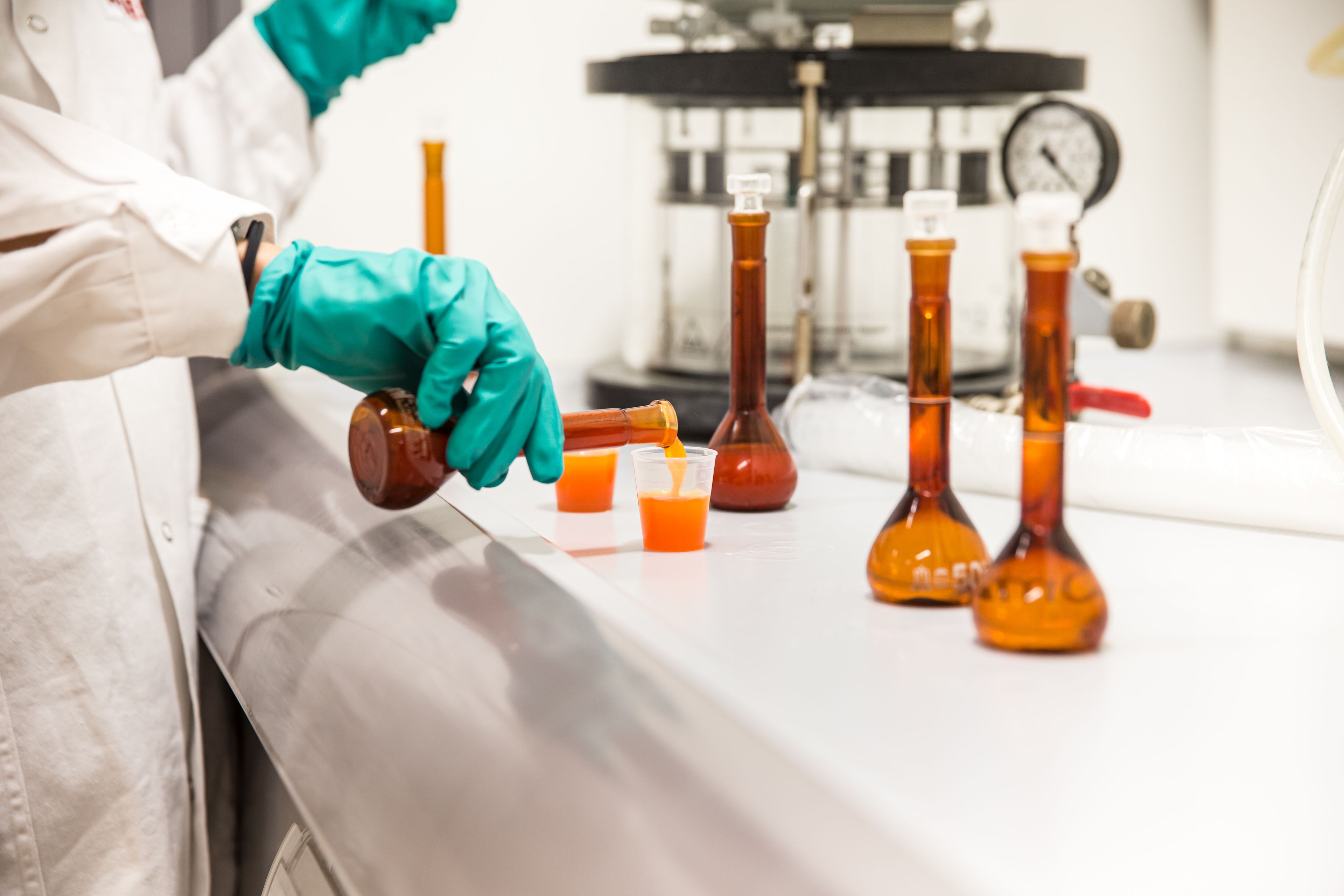 Mitarbeiterin im WIBERG Labor Freilassing in weißem Kittel und grünen Schutzhandschuhen gießt eine orange Flüssigkeit aus einem braunen Rundkolben in einen transparenten Behälter; auf der Arbeitsfläche stehen weitere Laborgläser, im Hintergrund unscharfe Laborumgebung.