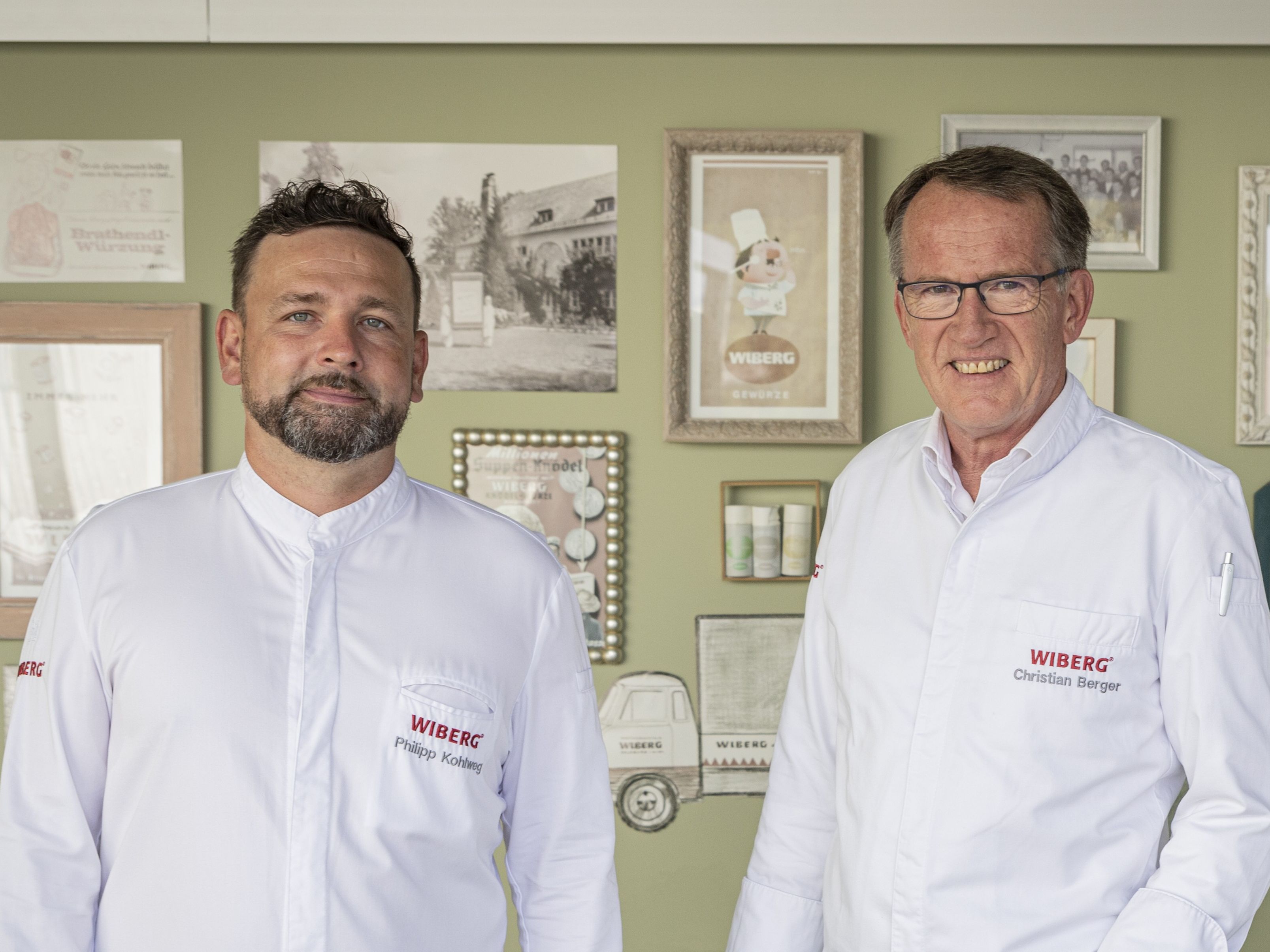 Etwas näher aufgenommenes Portrait von Philipp Kohlweg und Christian Berger vom WIBERG Team Inspiration in weißen Kochjacken, vor einer hellgrünen Wand mit Retro‑Werbemotiven und historischen Schwarzweiß‑Fotos aus der WIBERG Geschichte.