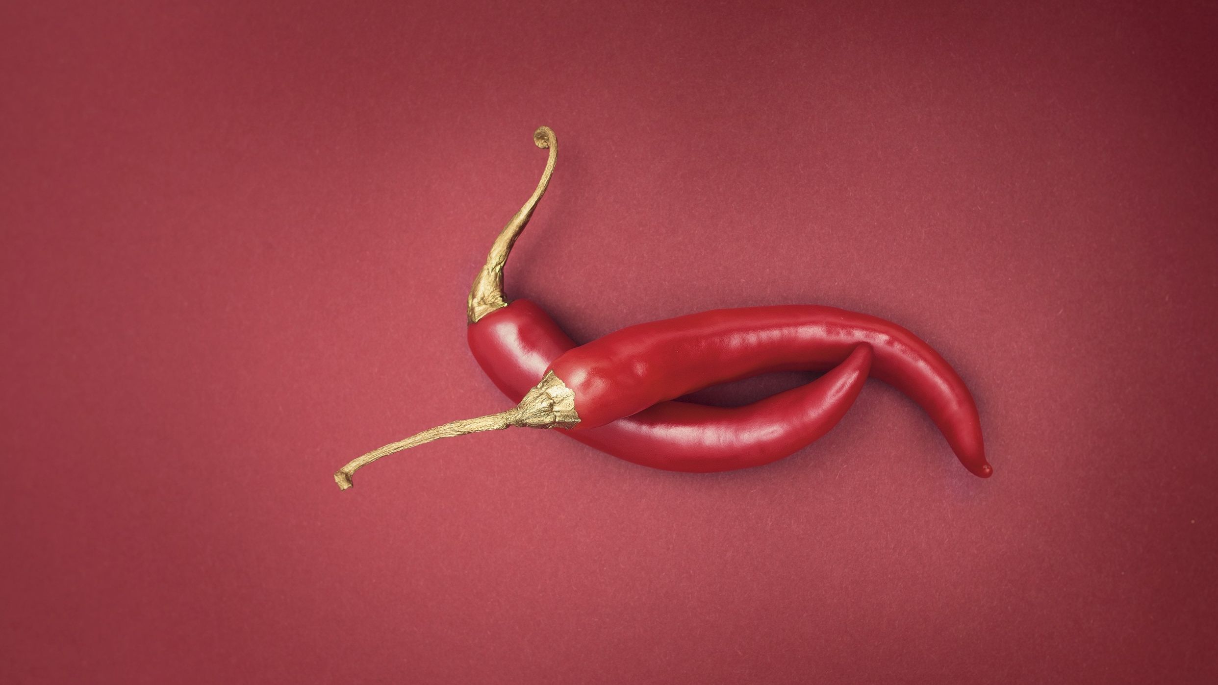 Von oben fotografierte zwei rote Chilischoten auf einem dunkelroten Hintergrund. Die Stiele der Chilischoten sind golden eingefärbt und sorgen für einen stimmungsvollen Farbkontrast.