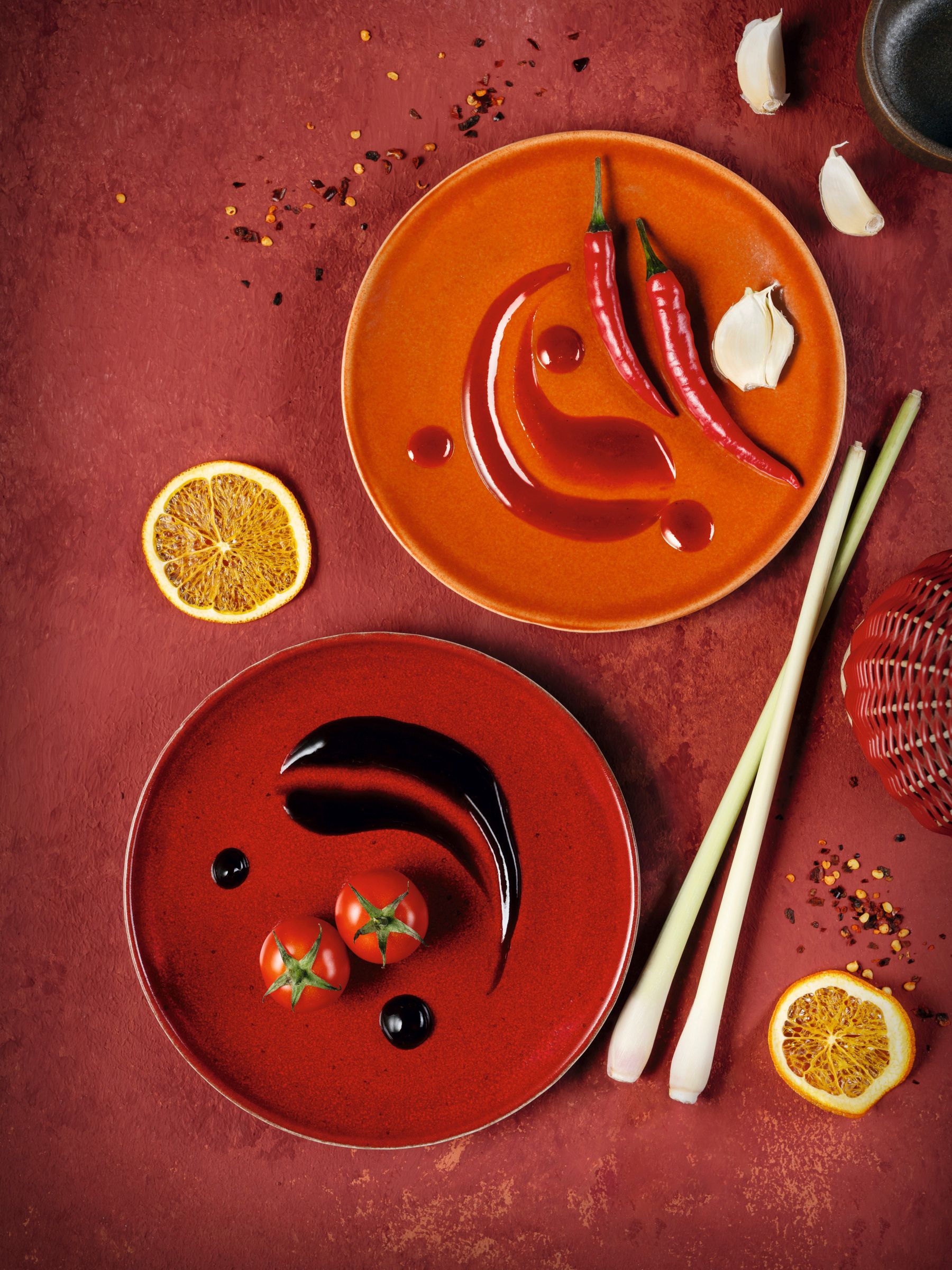 Zwei Teller mit WIBERG Würzsaucen: rote Spicy Chili mit Chilis und Knoblauch sowie dunkelbraune Tasty Umami mit Tomaten – moderne Draufsicht in kräftigem Rotton für Profiköche.