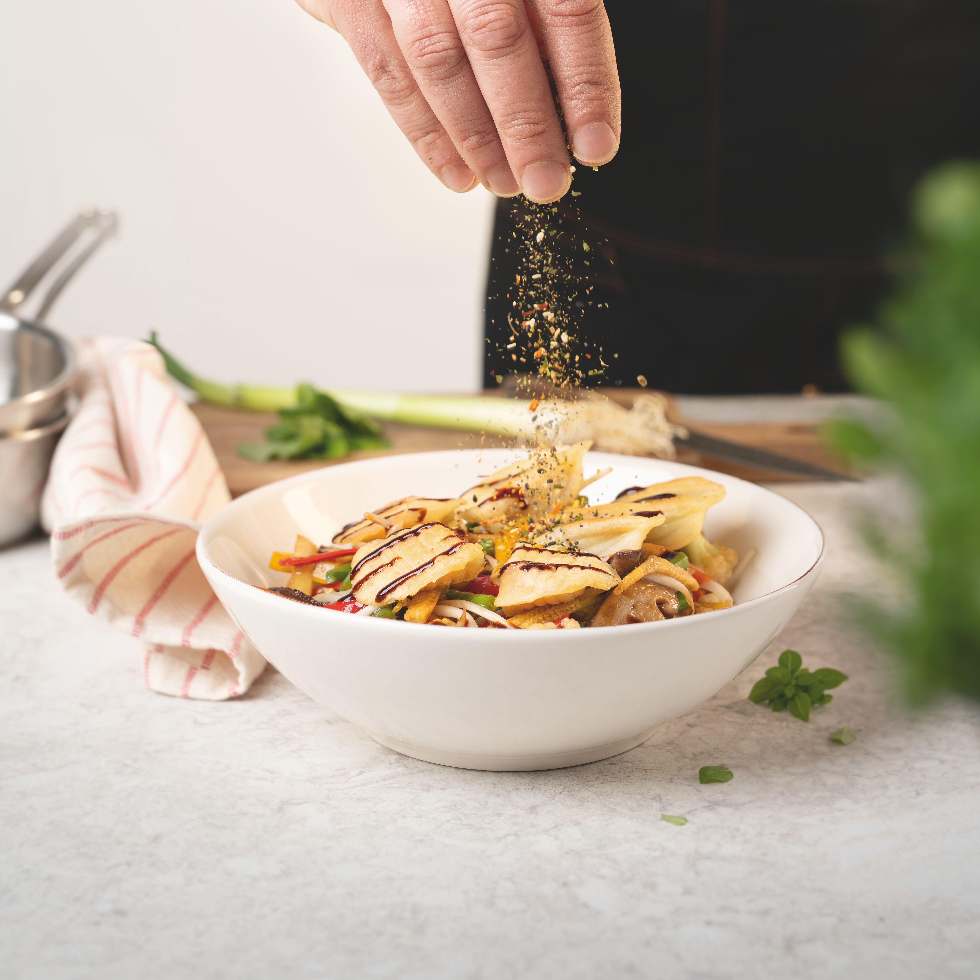 Hand eines Profikochs streut eine bunte WIBERG Gewürzmischung über dekorativ angerichtete Teigtaschen mit Gemüse; die Gewürze sind in der Luft im Streuvorgang sichtbar.