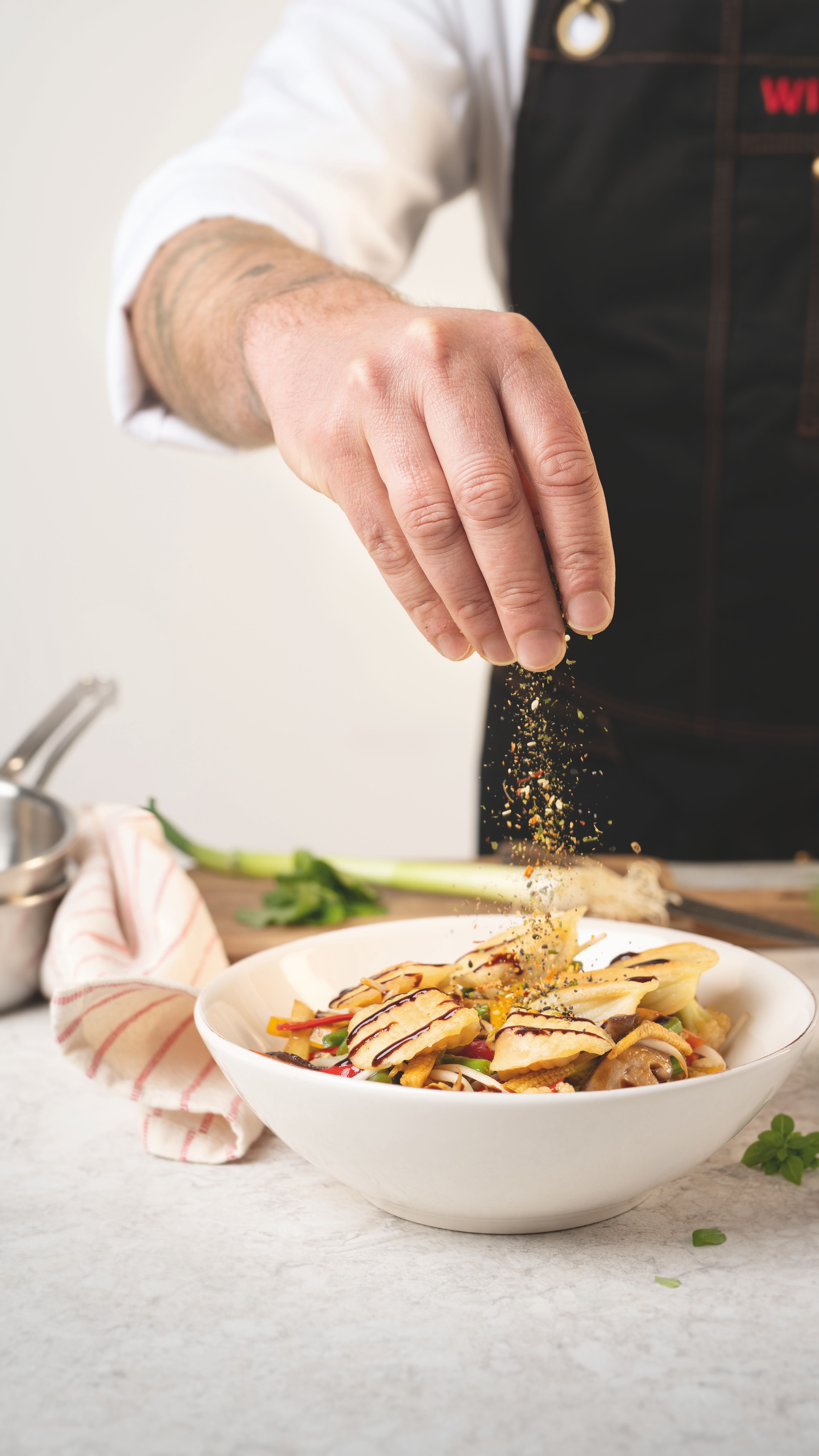 Hand eines Profikochs streut eine bunte WIBERG Gewürzmischung über dekorativ angerichtete Teigtaschen mit Gemüse; die Gewürze sind in der Luft im Streuvorgang sichtbar.