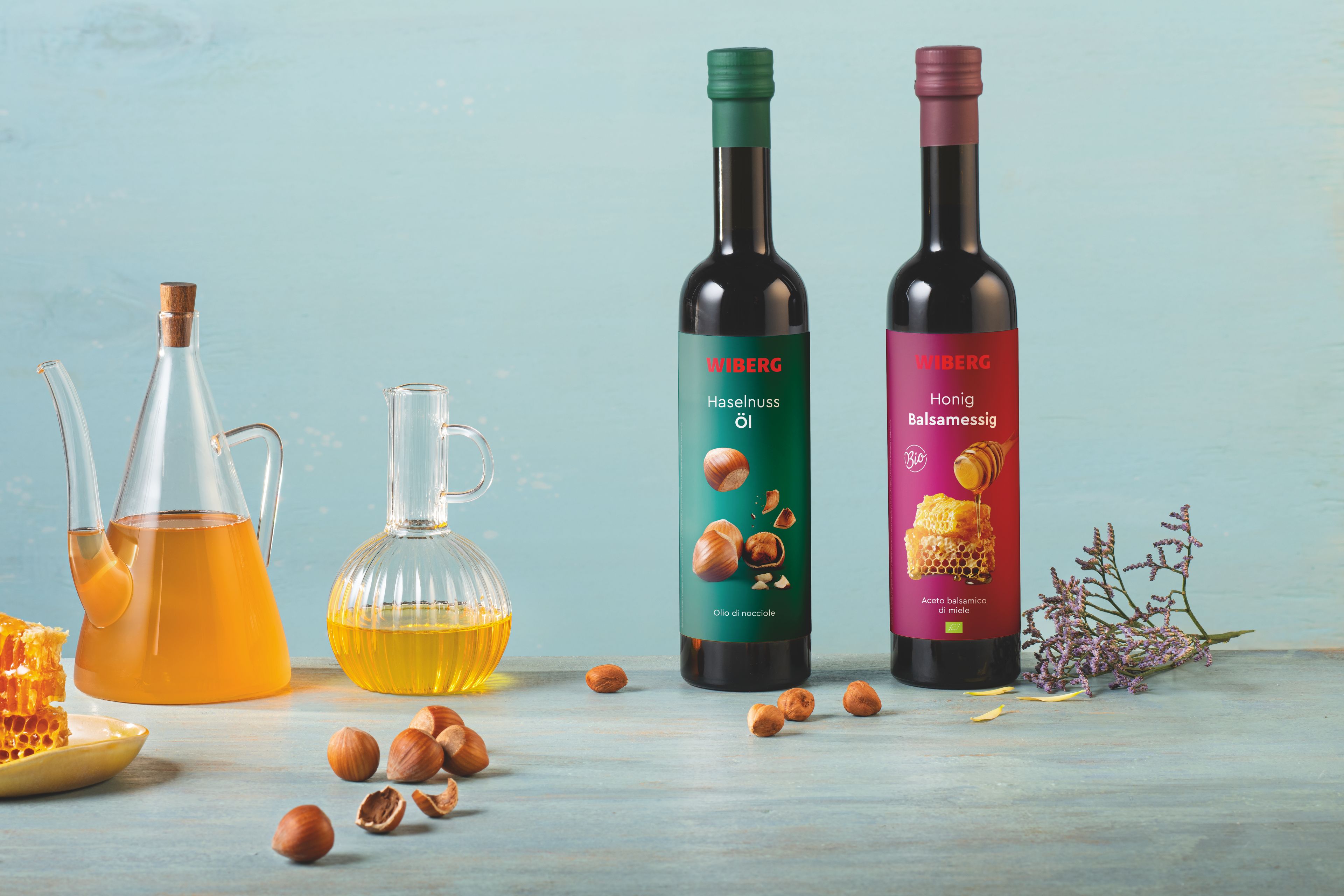 Von vorne fotografierte WIBERG Produkte auf pastellblauem Hintergrund: links Honigwabe und Haselnüsse, dahinter Karaffen mit WIBERG BIO Honig Balsamessig und Haselnuss Öl; rechts zwei 500 ml Flaschen mit grünem Haselnuss Öl Etikett und rotem BIO Honig Balsamessig Etikett.