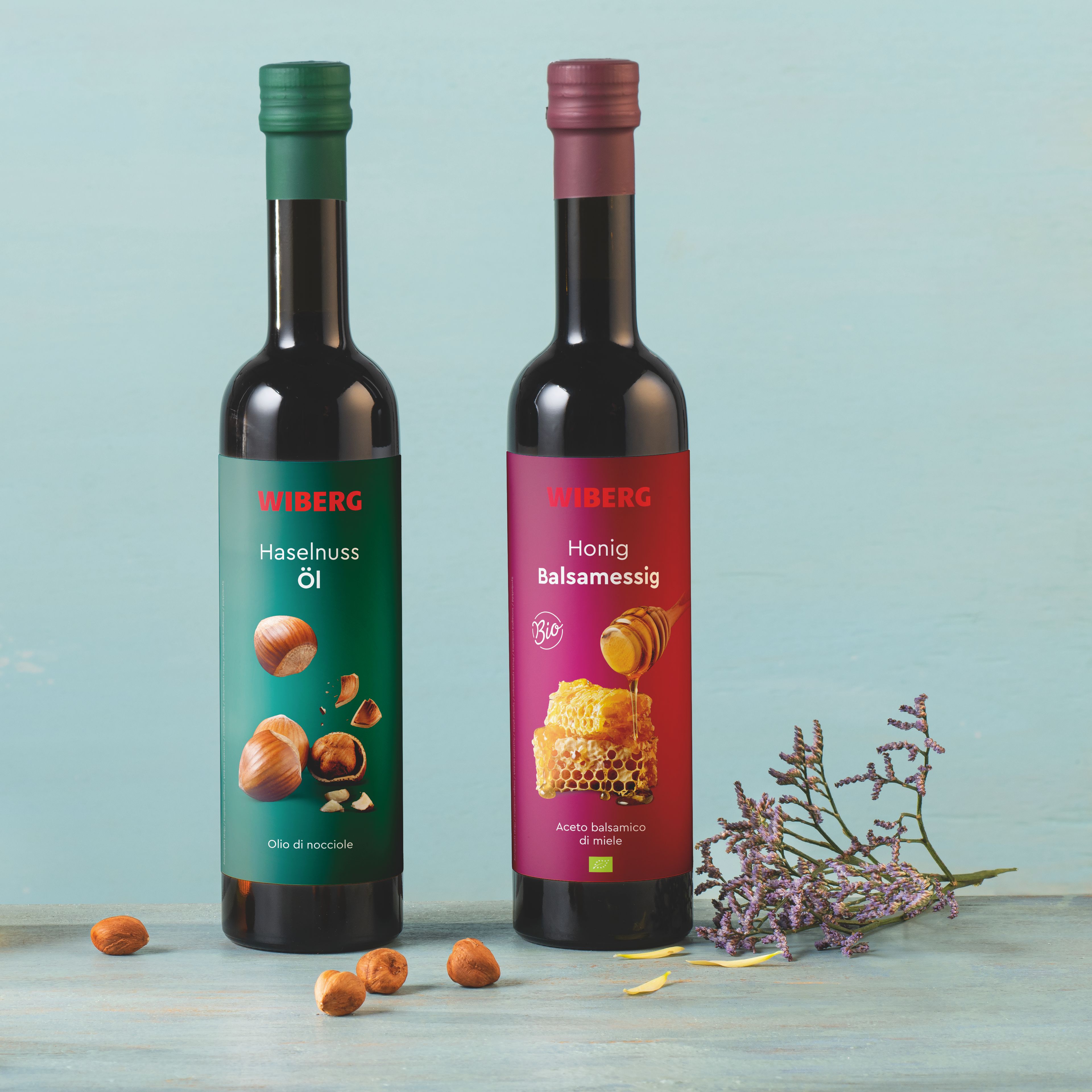 Von vorne fotografierte WIBERG Produkte auf pastellblauem Hintergrund: links Honigwabe und Haselnüsse, dahinter Karaffen mit WIBERG BIO Honig Balsamessig und Haselnuss Öl; rechts zwei 500 ml Flaschen mit grünem Haselnuss Öl Etikett und rotem BIO Honig Balsamessig Etikett.