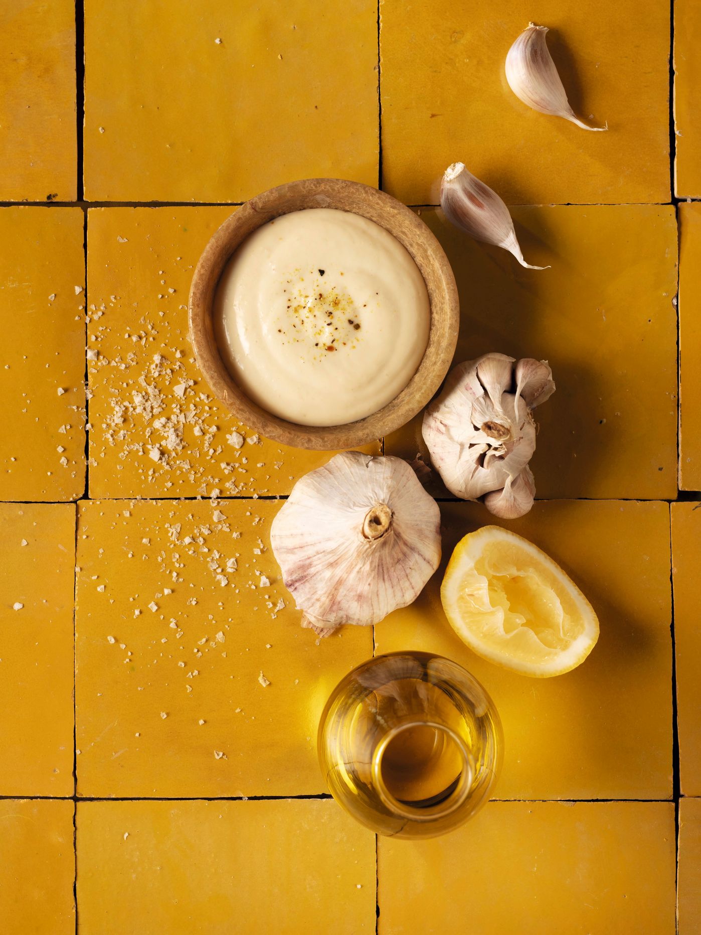 WIBERG Gourmetsauce Aioli in einer kleinen Schüssel, umgeben von Knoblauch, Zitrone, Meersalz und nativem Olivenöl – Draufsicht im warmen Gelbton, ideal für Profiköche.