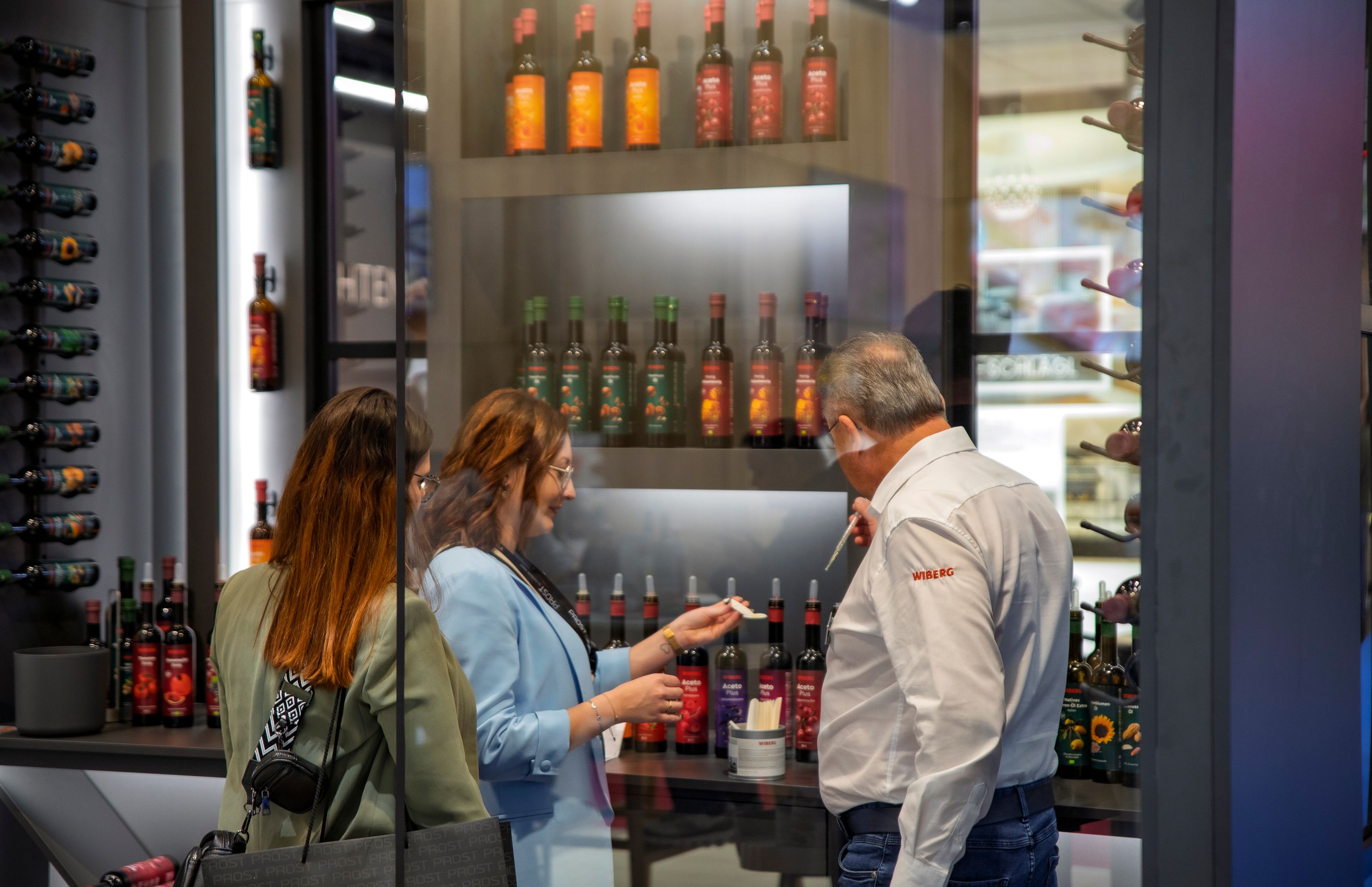 Ein Vertriebsmitarbeiter präsentiert zwei Besucherinnen am WIBERG Messestand die WIBERG Premium-Essige und Premium-Öle zur Verkostung. Die Flüssigprodukte mit neu designten Etiketten stehen in einem edel gestalteten Showroom und werden hochwertig inszeniert.