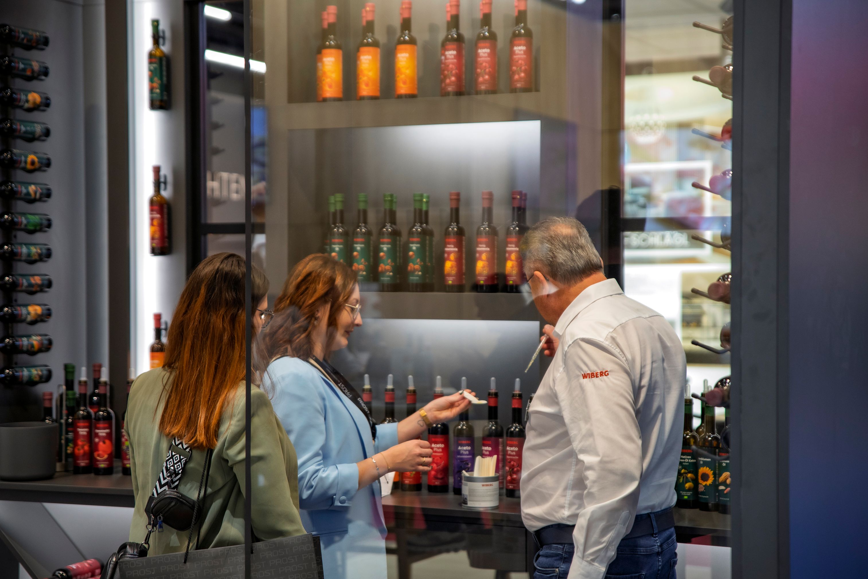 Ein Vertriebsmitarbeiter präsentiert zwei Besucherinnen am WIBERG Messestand die WIBERG Premium-Essige und Premium-Öle zur Verkostung. Die Flüssigprodukte mit neu designten Etiketten stehen in einem edel gestalteten Showroom und werden hochwertig inszeniert.