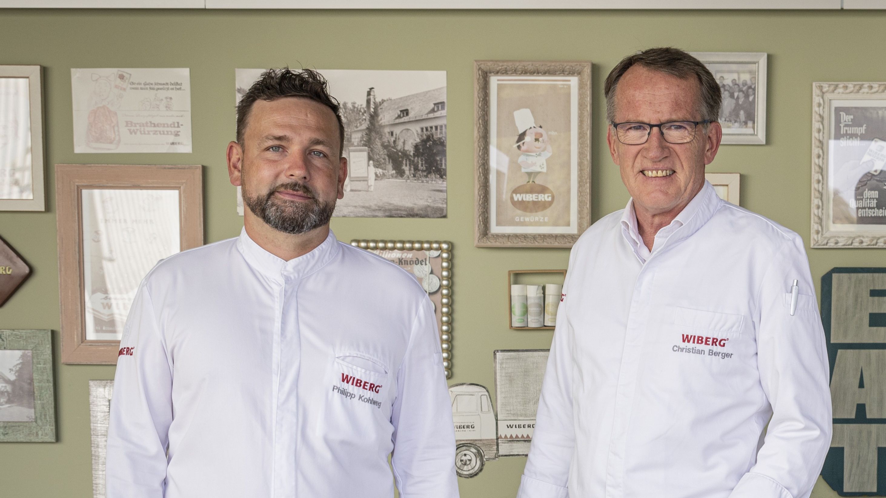 Philipp Kohlweg und Christian Berger vom WIBERG Team Inspiration in weißen Kochjacken vor einer hellgrünen Wand mit Retro‑Werbemotiven und historischen Schwarzweiß‑Fotos aus der WIBERG Geschichte.