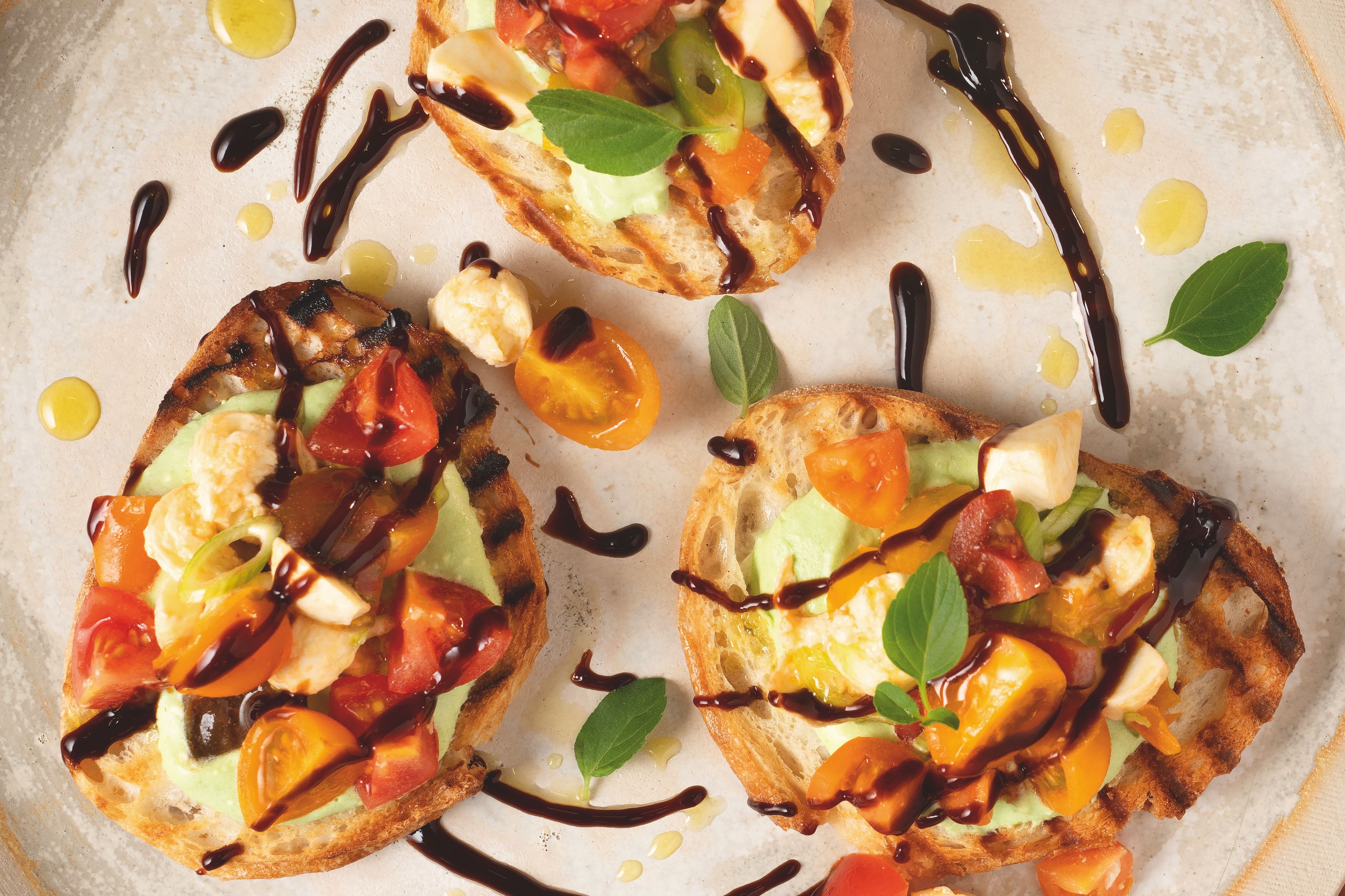 Von oben fotografierter Teller mit drei Bruschetta‑Brötchen, belegt mit rot‑ und dunkelgelben Tomaten, verfeinert mit WIBERG Würzsauce Tasty Umami und frischem Basilikum, auf rostrotem Hintergrund.