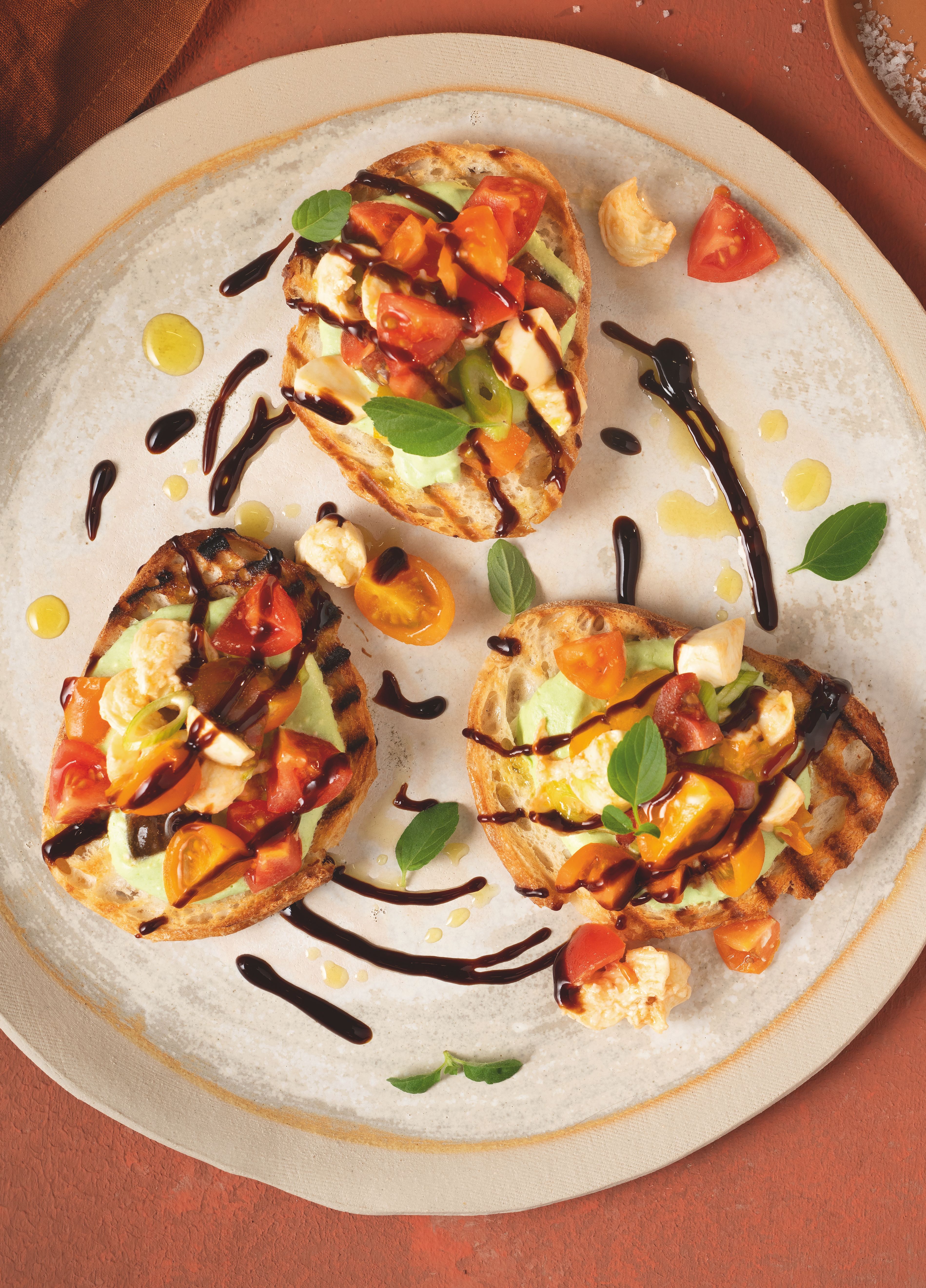 Von oben fotografierter Teller mit drei Bruschetta‑Brötchen, belegt mit rot‑ und dunkelgelben Tomaten, verfeinert mit WIBERG Würzsauce Tasty Umami und frischem Basilikum, auf rostrotem Hintergrund.