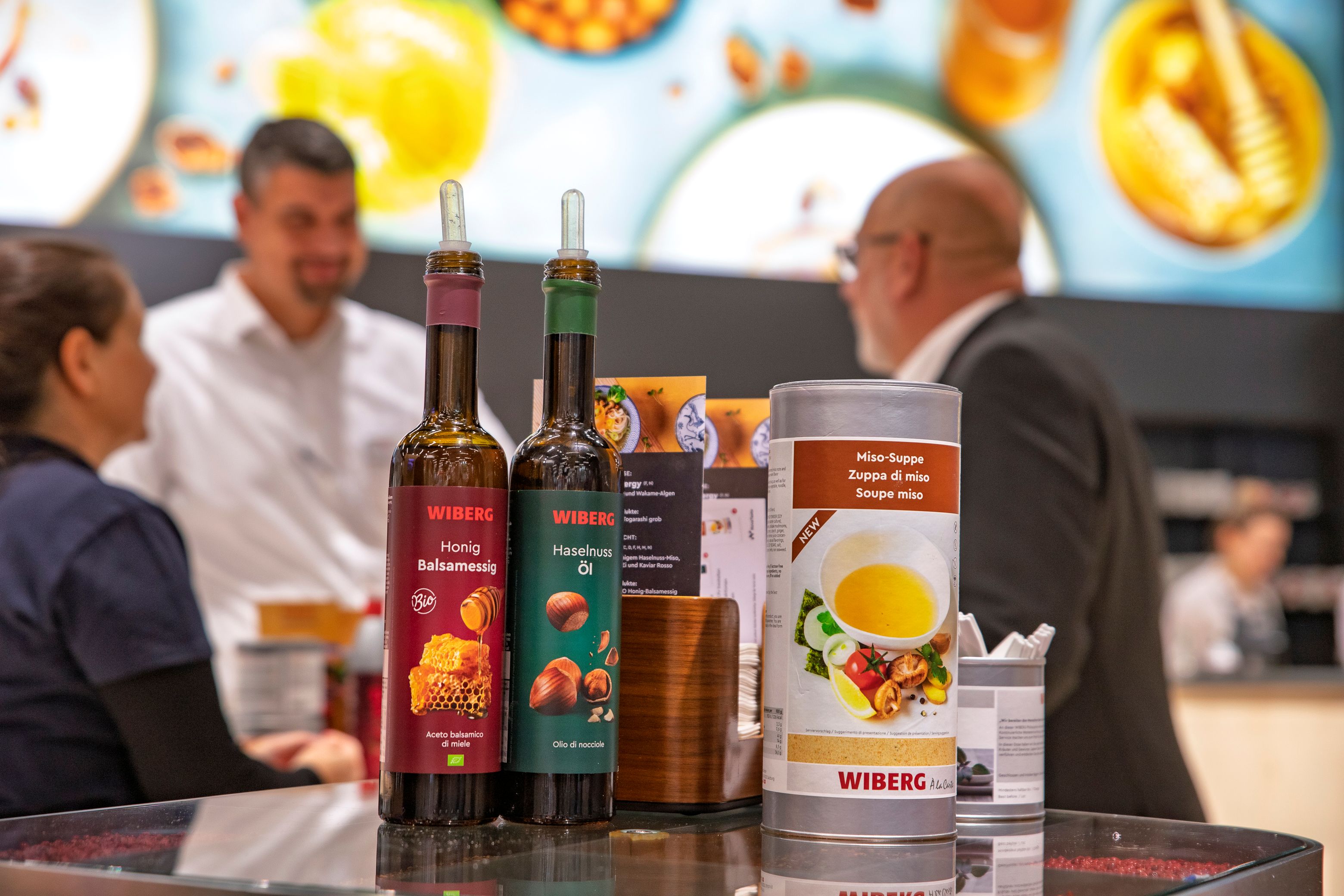 Im Vordergrund stehen die WIBERG Neuprodukte BIO Honig Balsamessig, Haselnuss Öl mit neu designten Etiketten sowie Miso Suppe auf einem Hochtisch am WIBERG Messestand einer Gastromesse. 