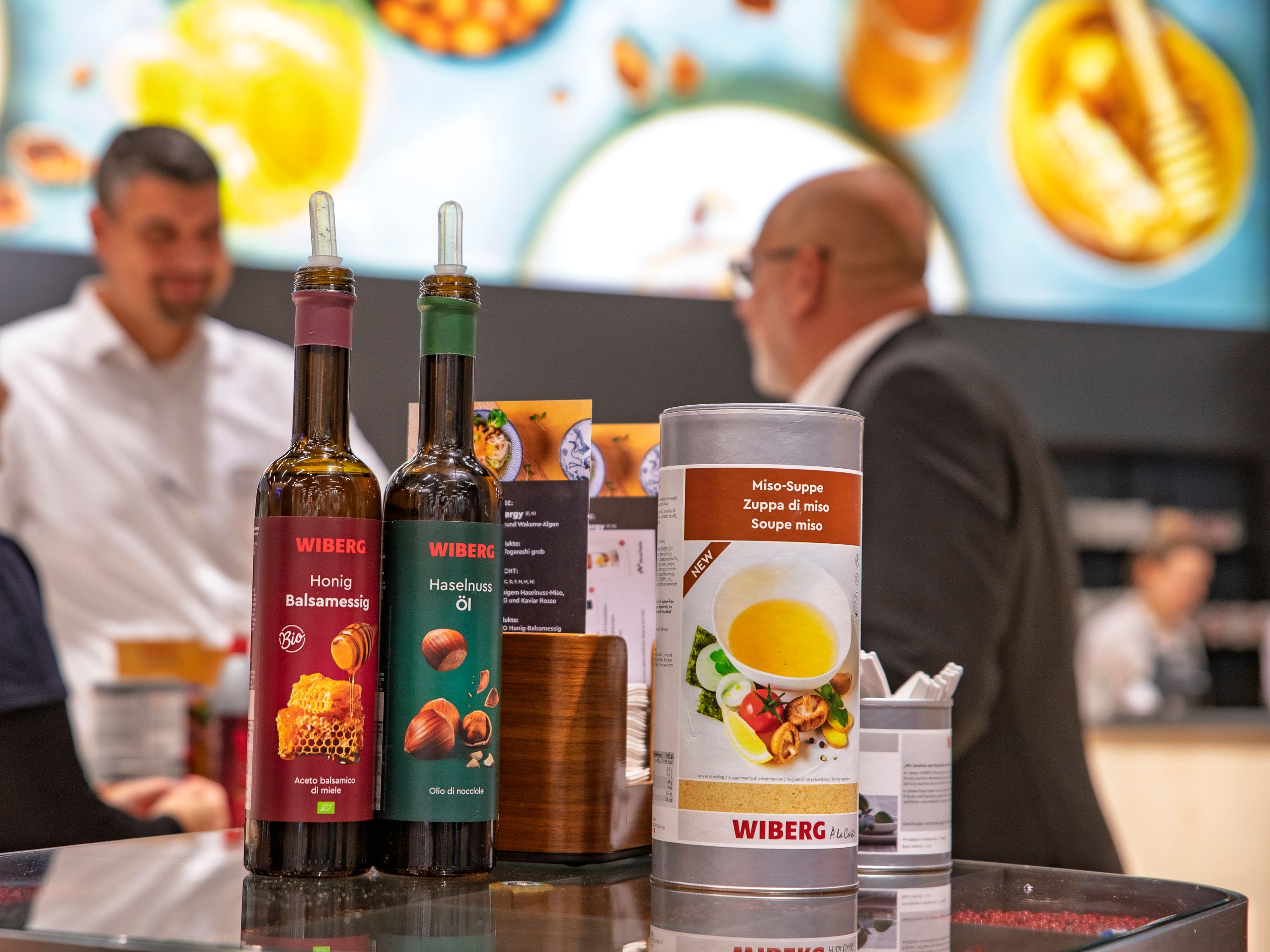 Im Vordergrund stehen die WIBERG Neuprodukte BIO Honig Balsamessig, Haselnuss Öl mit neu designten Etiketten sowie Miso Suppe auf einem Hochtisch am WIBERG Messestand einer Gastromesse. 
