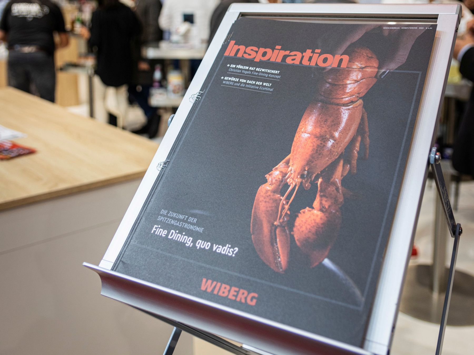Im Vordergrund liegt das WIBERG Kundenmagazin „Inspiration“ mit schwarzem Titelblatt und Hummermotiv als Fine‑Dining‑Ausgabe auf einem Zeitschriftenständer am WIBERG Messestand einer Gastronomiemesse. Im unscharfen Hintergrund sind Besucher am Messestand zu sehen.