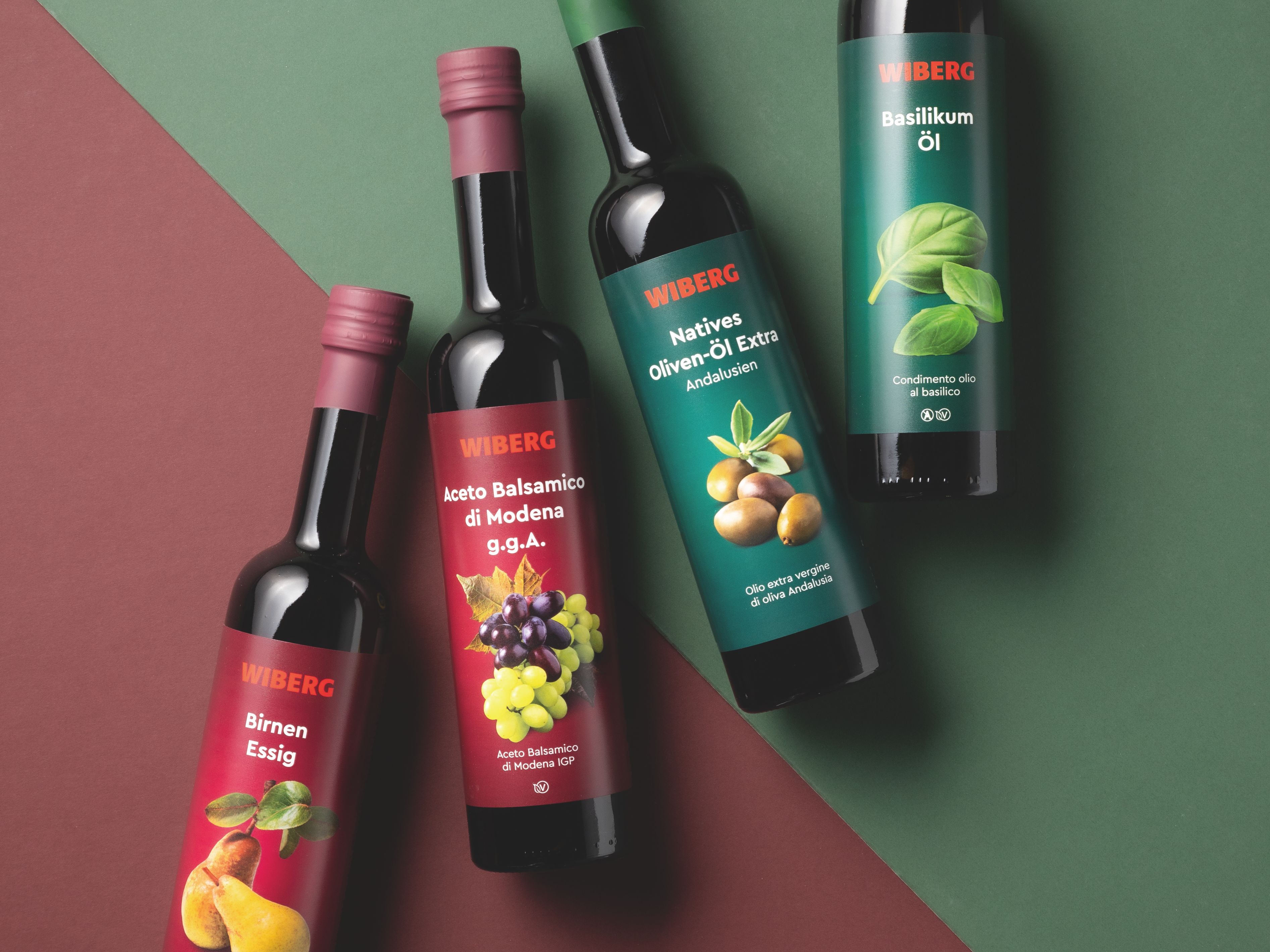 WIBERG Birnen Essig und WIBERG Aceto Balsamico di Modena g.g.A. mit dunkelroten Etiketten sowie WIBERG Natives Oliven Öl Extra Andalusien und WIBERG Basilikum Öl mit dunkelgrünen Etiketten in dunklen 500 ml Flaschen auf diagonal geteiltem Hintergrund in Dunkelrot und Dunkelgrün.