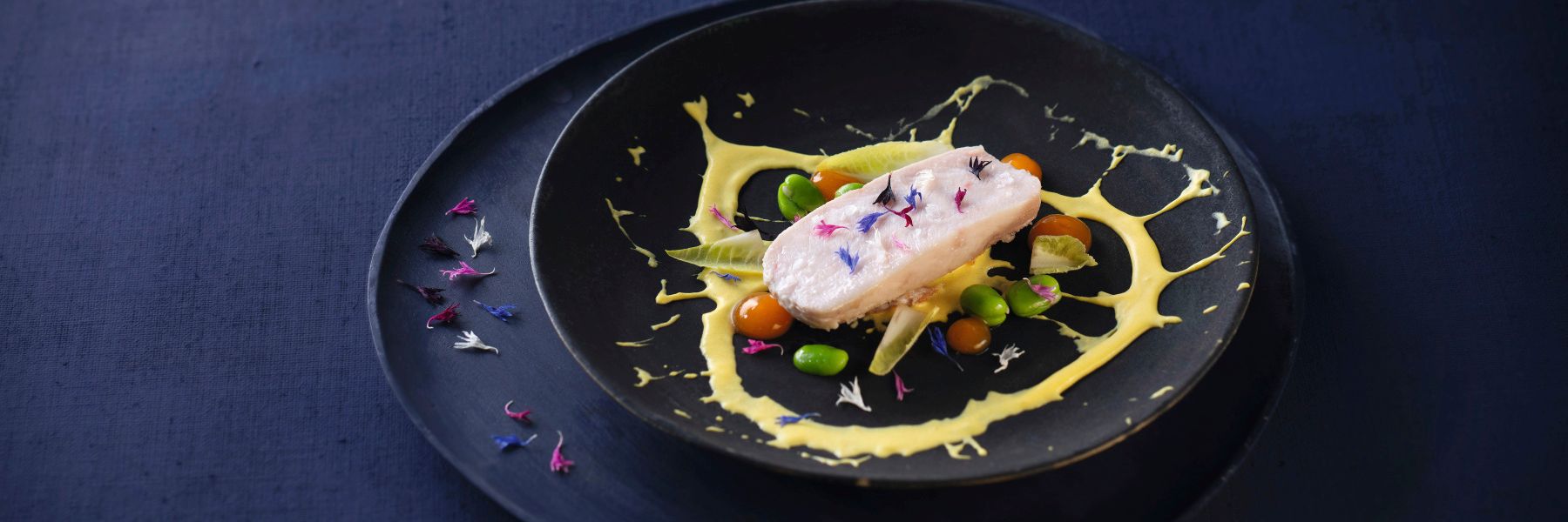 Kaninchenrücken mit Ackerbohnen, Safran und Kornblumenblüten dekorativ auf dunklem Teller vor dunkelviolettem Hintergrund – farbintensives Gourmetgericht mit WIBERG Exquisite Produkten für Profiköche.