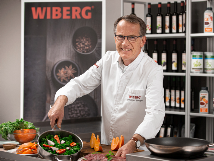 Christian Berger vom WIBERG Team Inspiration in weißer Kochjacke vor einem Regal mit WIBERG Produkten. Er präsentiert eine Pfanne mit frischen Zutaten.