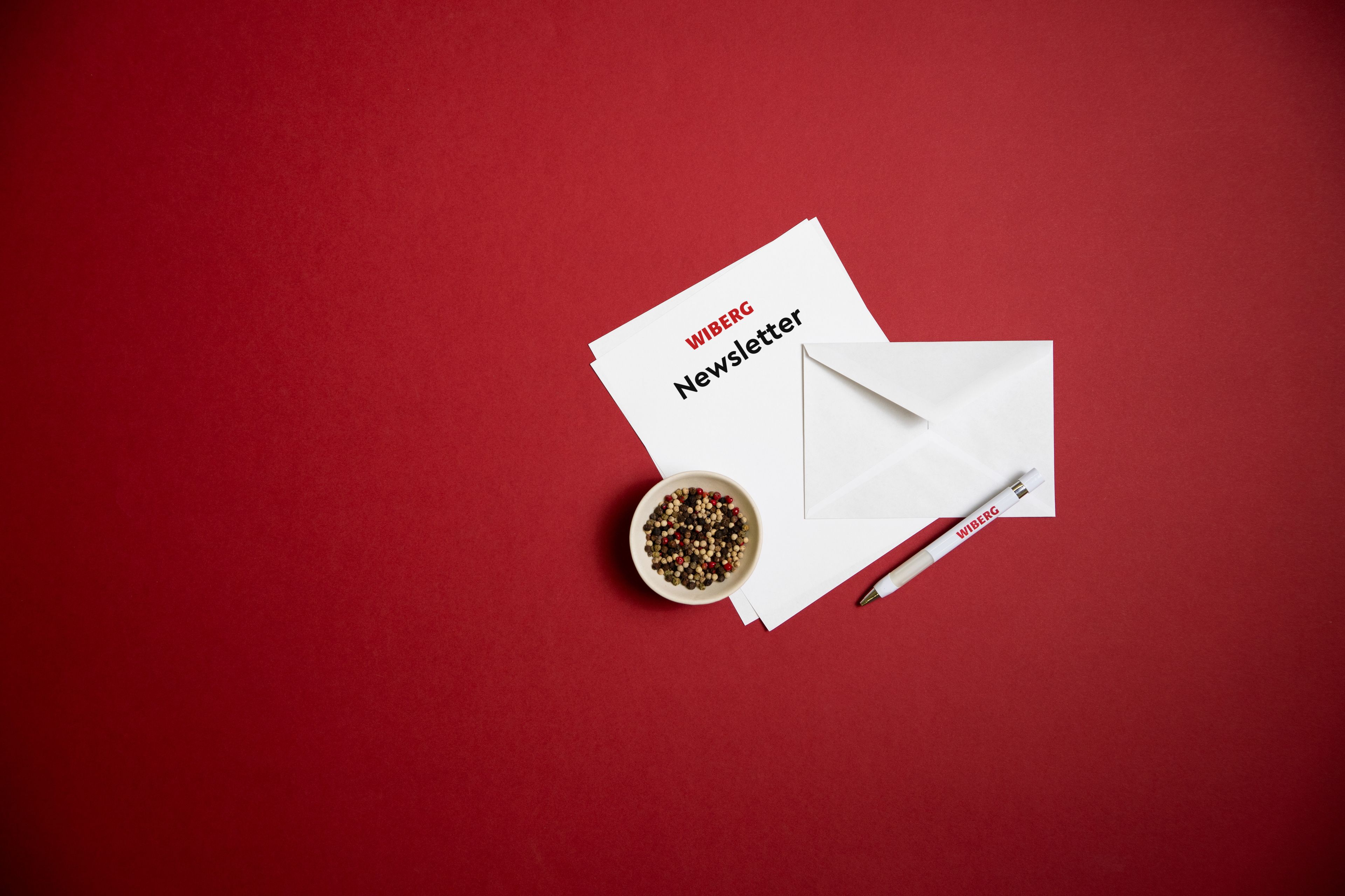 Spotlight auf einen weißen Brief mit rotem WIBERG Logo und der Aufschrift „Newsletter“, dazu ein weißes Kuvert, ein WIBERG Kugelschreiber und eine kleine Schüssel bunter Pfefferkörner vor dunkelrotem Hintergrund.