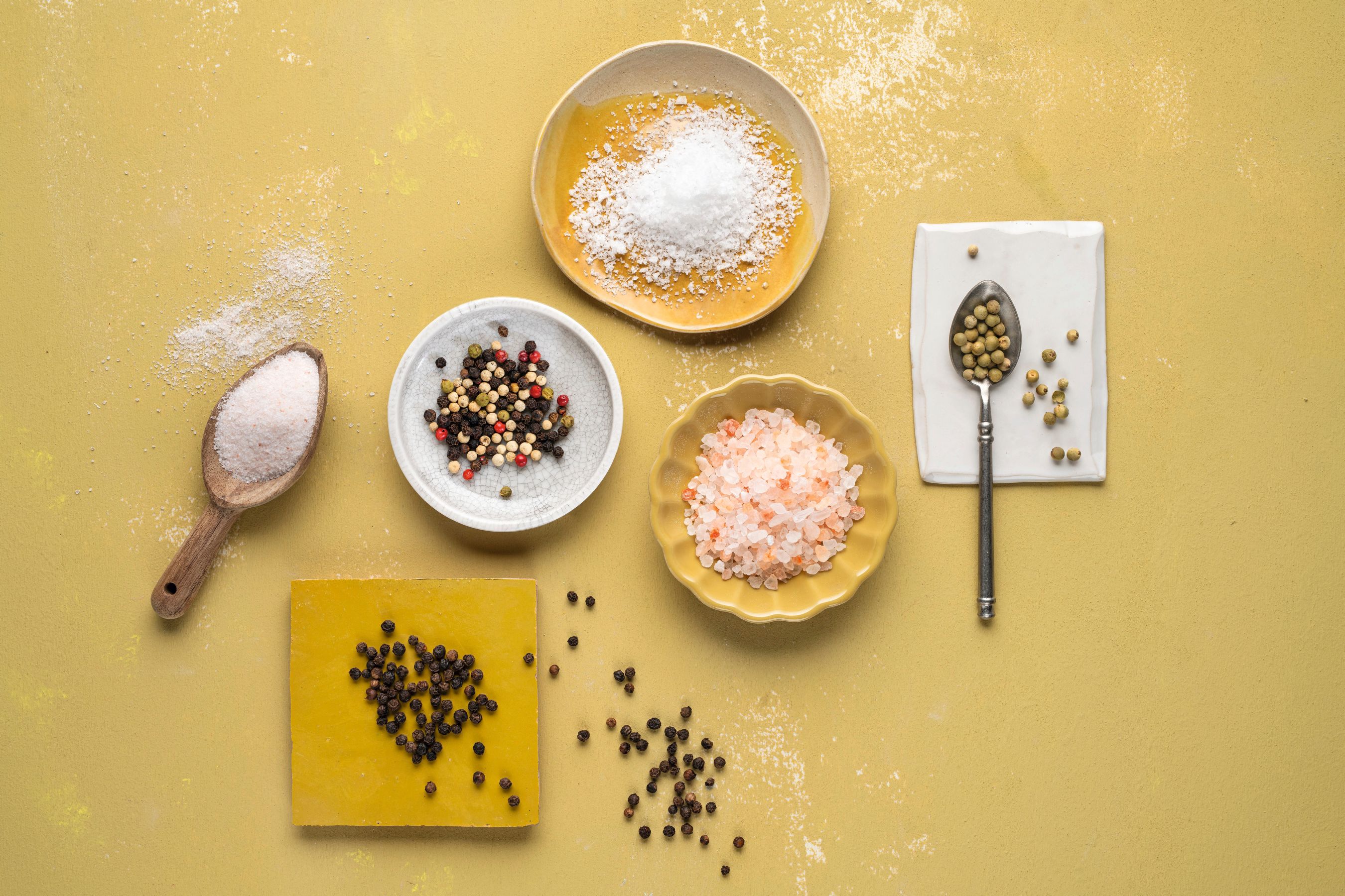Von oben aufgenommenes WIBERG Bild mit verschiedenen Pfeffer- und Salzsorten. Rosa Ursalz, weißes Meersalz und bunte Pfeffersorten liegen locker arrangiert auf weißen und gelben Tellern und erzeugen ein freundliches, stimmungsvolles Gesamtbild.