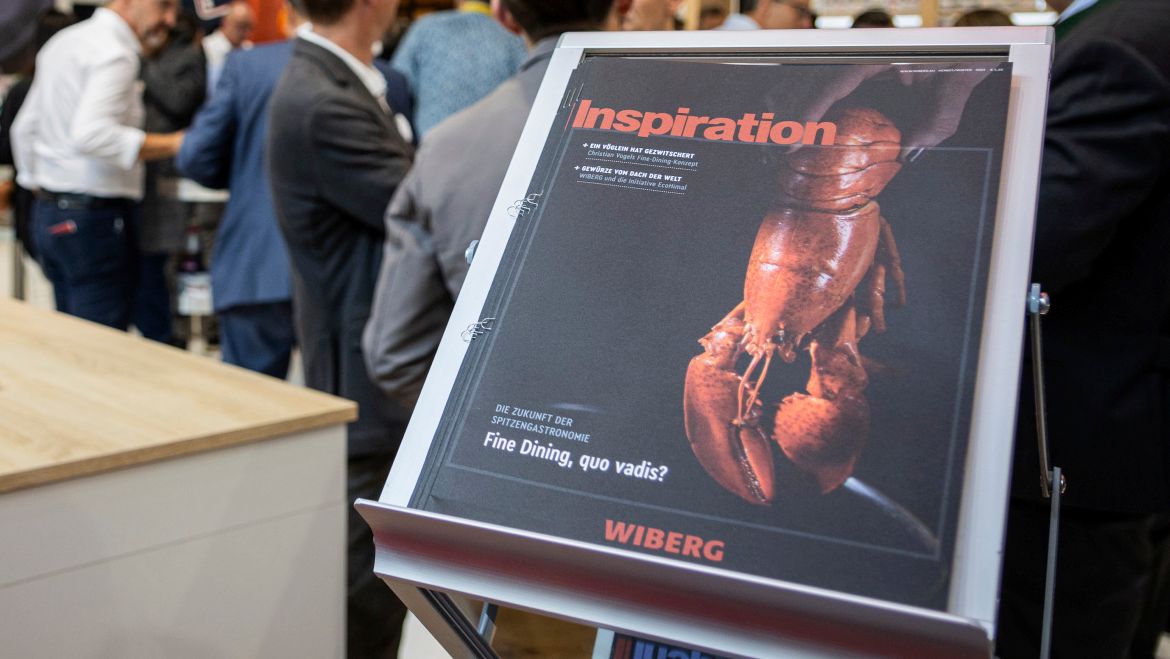 Im Querformat aufgenommenes Bild vom WIBERG Kundenmagazin „Inspiration“ mit schwarzem Titelblatt und Hummermotiv als Fine Dining Ausgabe. Das Magazin liegt auf einem Zeitschriftenständer am WIBERG Messestand einer Gastronomiemesse, im Hintergrund unscharf Besucher auf dem gut frequentierten Stand.