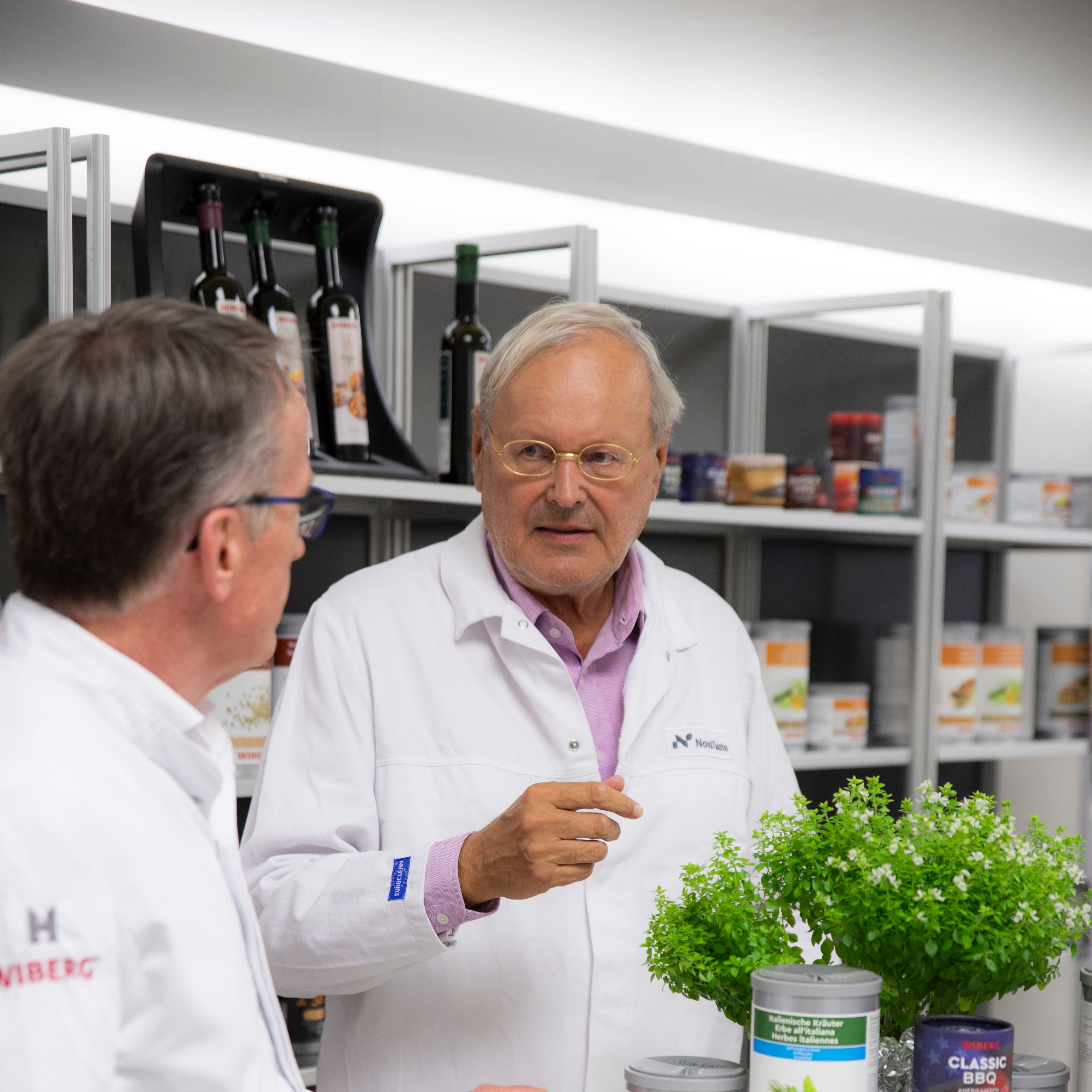 Christian Berger und  Prof. Dr. med. Markus M. Metka in weißen Arbeitskitteln im Gespräch vor frischen Kräutern und WIBERG-Gewürzprodukten