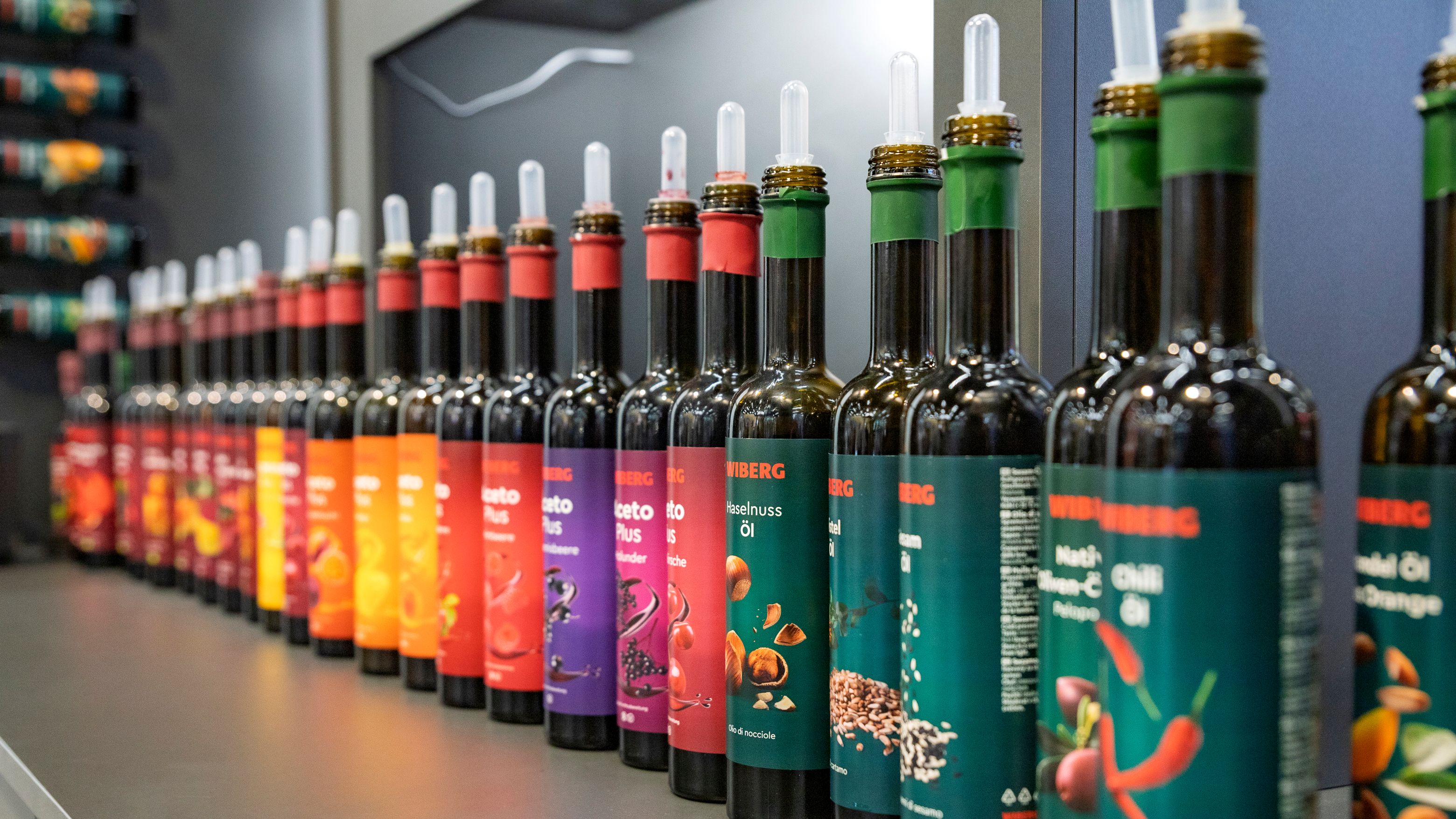 Reihe von etwa 20 dunklen 500 ml Flaschen der WIBERG Premium-Essige, Premium-Öle sowie AcetoPlus Fruchtessige mit farbenfrohen neuen Etiketten auf einer Gastro-Messe präsentiert.