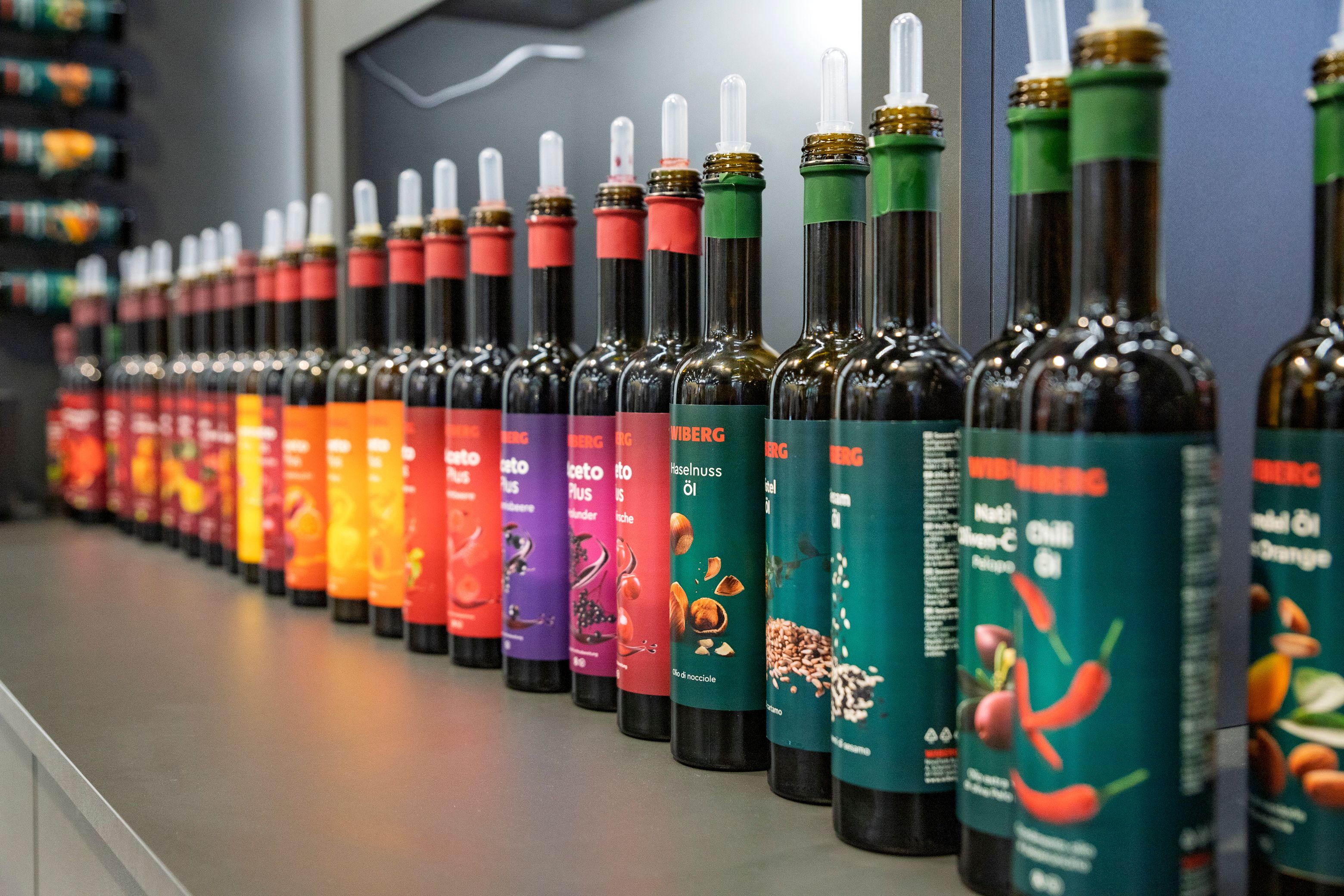 Reihe von etwa 20 dunklen 500 ml Flaschen der WIBERG Premium-Essige, Premium-Öle sowie AcetoPlus Fruchtessige mit farbenfrohen neuen Etiketten auf einer Gastro-Messe präsentiert.