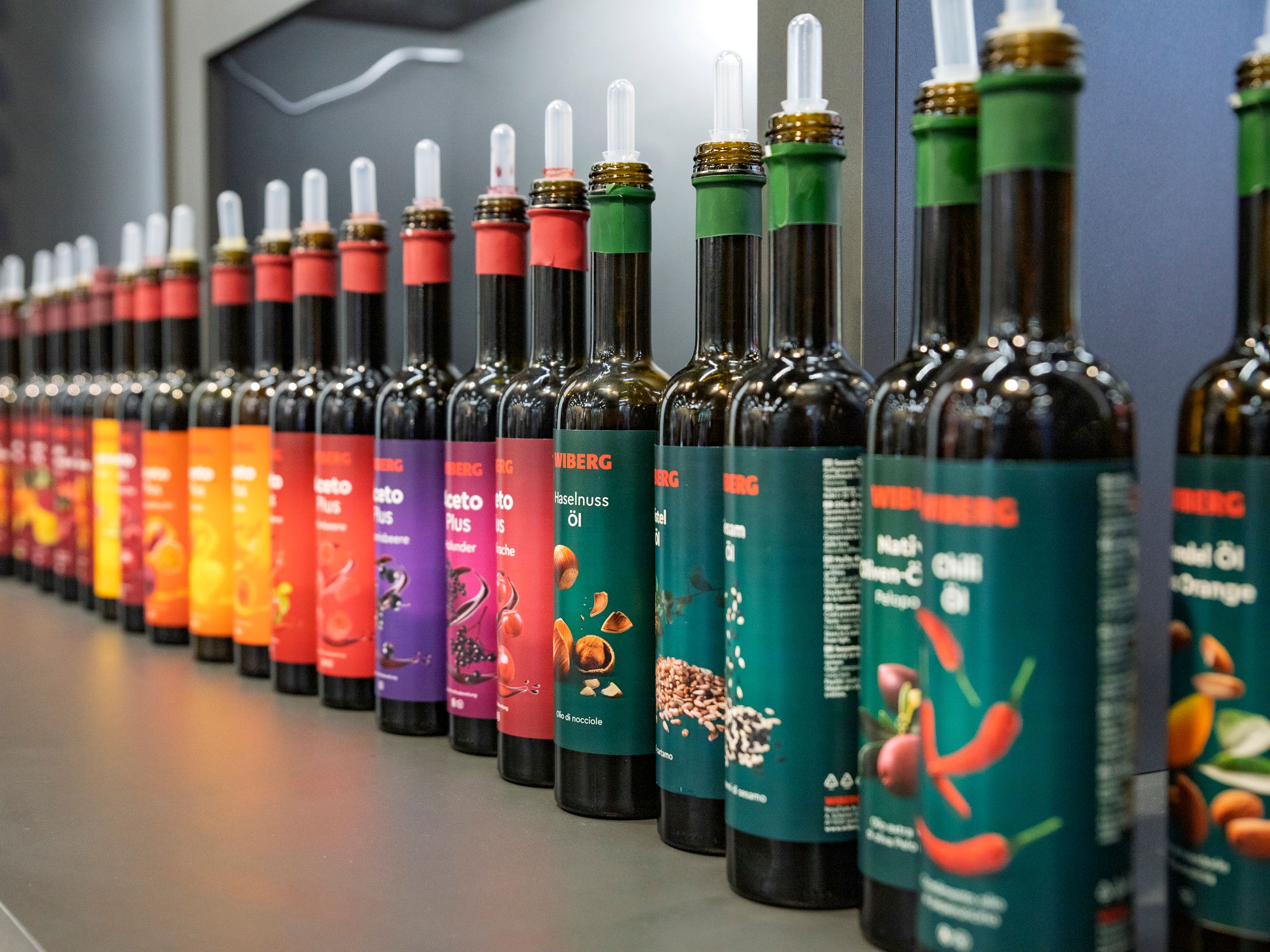 Reihe von etwa 20 dunklen 500 ml Flaschen der WIBERG Premium-Essige, Premium-Öle sowie AcetoPlus Fruchtessige mit farbenfrohen neuen Etiketten auf einer Gastro-Messe präsentiert.