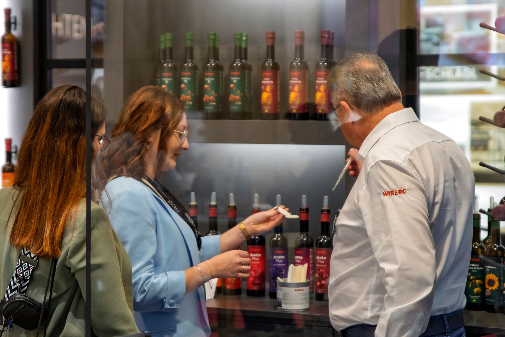 Ein Vertriebsmitarbeiter präsentiert zwei Besucherinnen am WIBERG Messestand die WIBERG Premium-Essige und Premium-Öle zur Verkostung. Die Flüssigprodukte mit neu designten Etiketten stehen in einem edel gestalteten Showroom und werden hochwertig inszeniert.