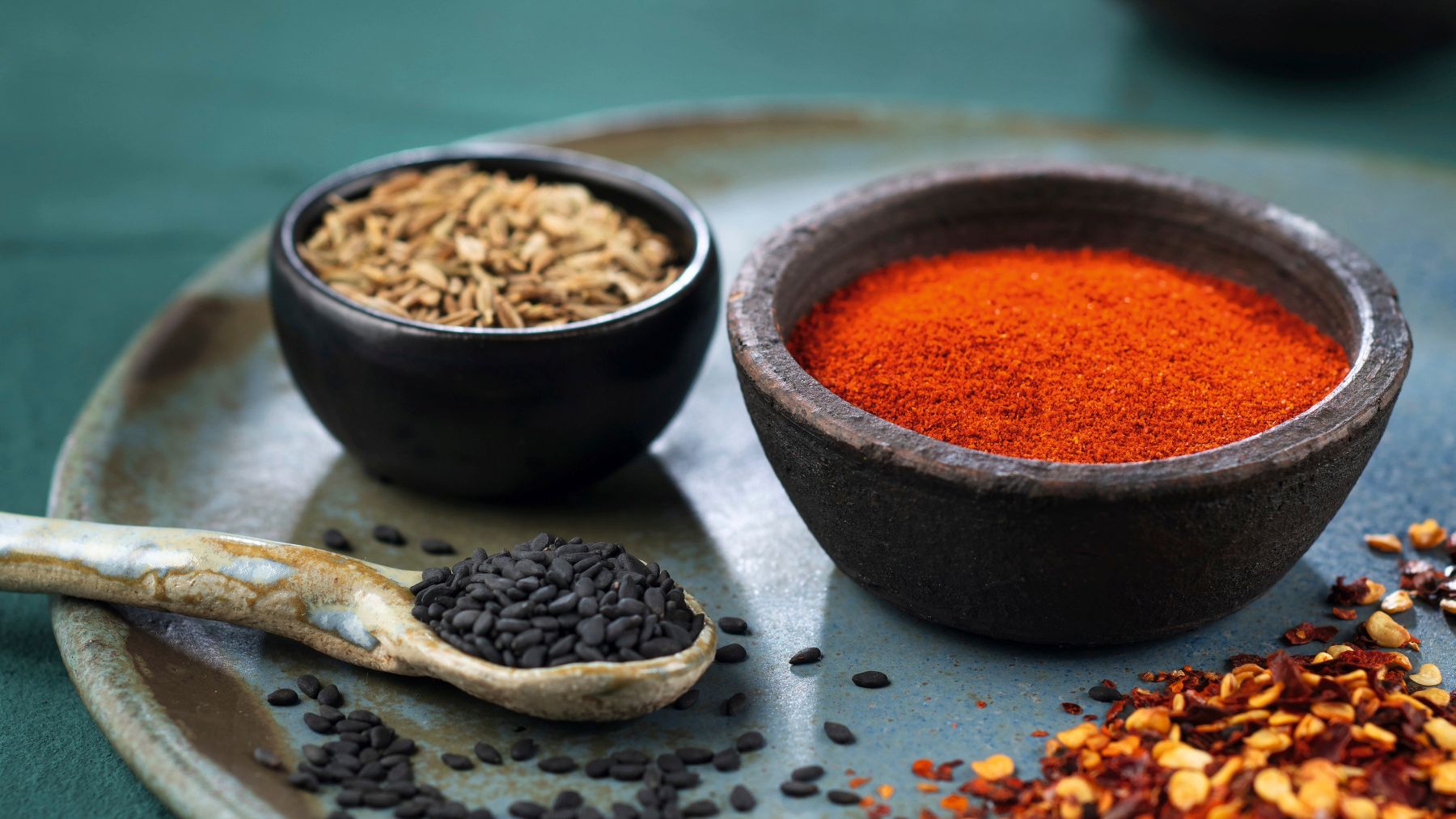 Von vorne fotografierte WIBERG Gewürze auf dunkelgrünem Hintergrund. Chili, schwarzer Sesam, Fenchel, Paprika und Curcuma liegen in Schüsseln und sind stimmungsvoll auf einem handgemachten Teller und Löffel verteilt.