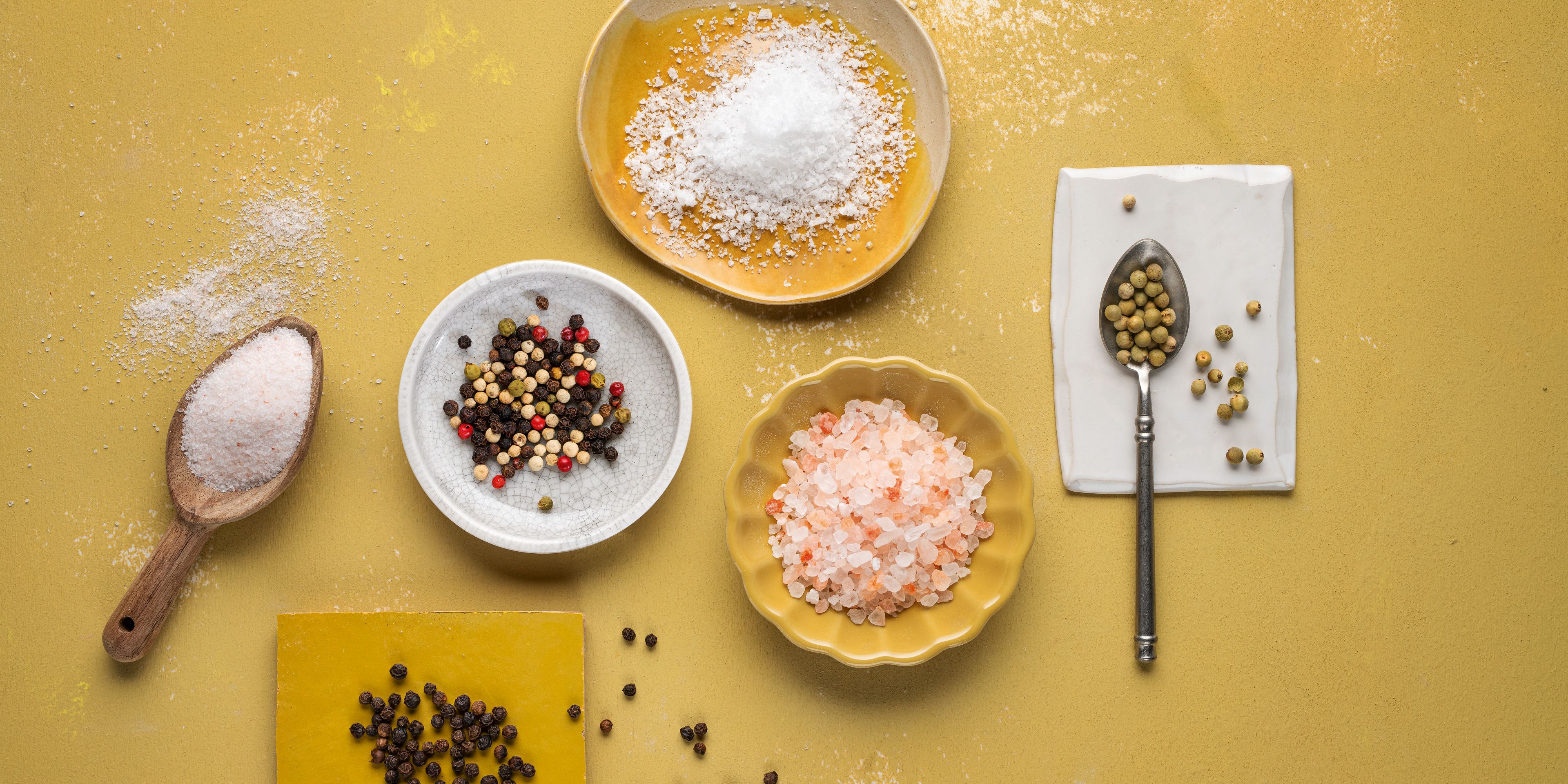 Von oben fotografierte WIBERG Pfeffer- und Salzsorten: Rosa Ursalz, weißes Meersalz und bunte Pfeffersorten locker auf weißen und gelben Tellern arrangiert auf hellem Hintergrund.