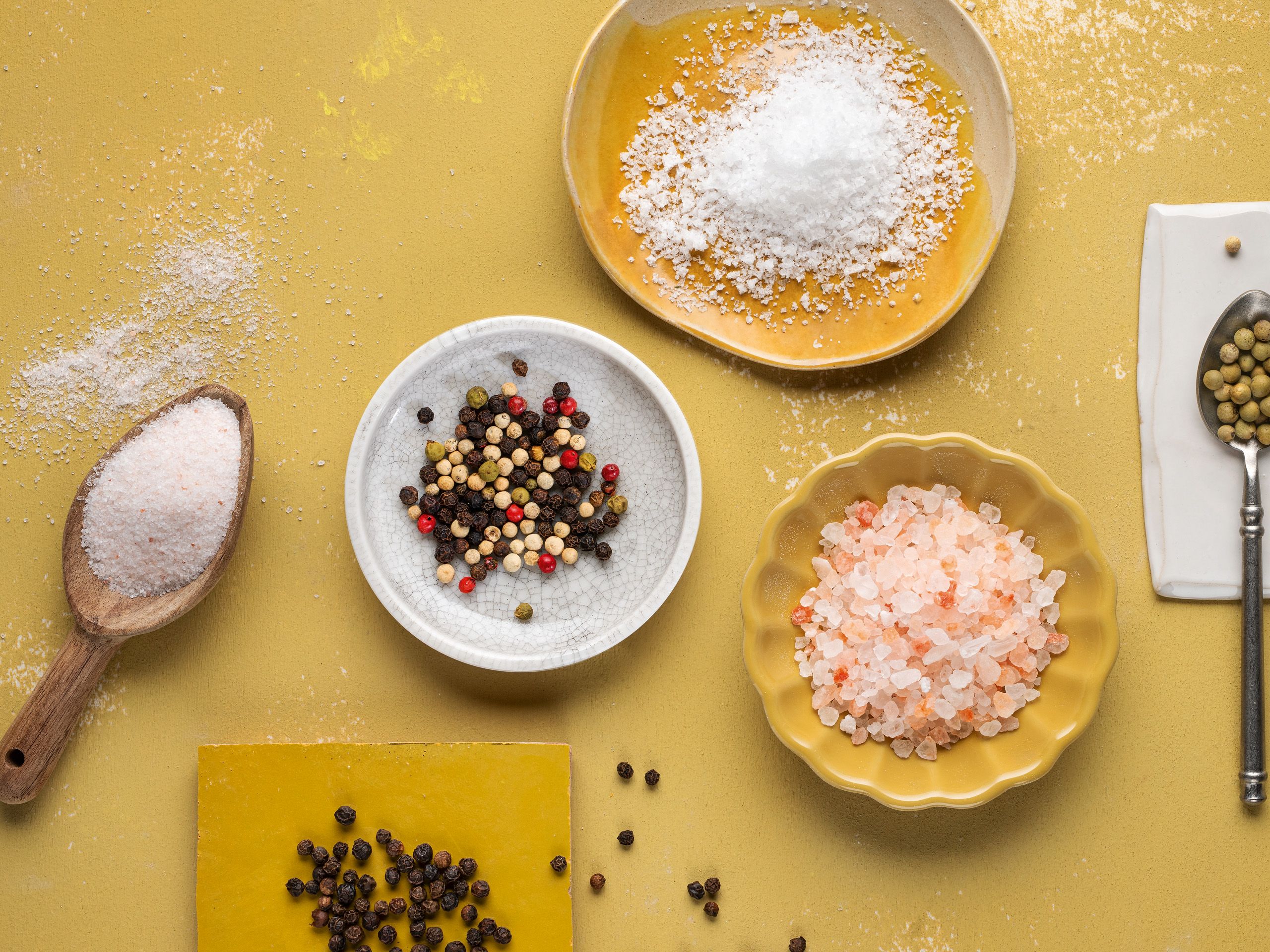 Von oben fotografierte WIBERG Pfeffer- und Salzsorten: Rosa Ursalz, weißes Meersalz und bunte Pfeffersorten locker auf weißen und gelben Tellern arrangiert auf hellem Hintergrund.