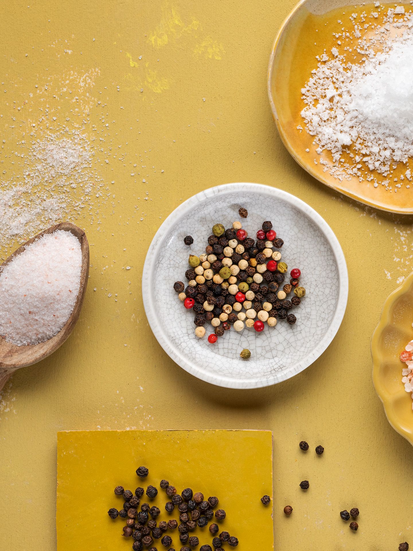 Von oben fotografierte WIBERG Pfeffer- und Salzsorten: Rosa Ursalz, weißes Meersalz und bunte Pfeffersorten locker auf weißen und gelben Tellern arrangiert auf hellem Hintergrund.