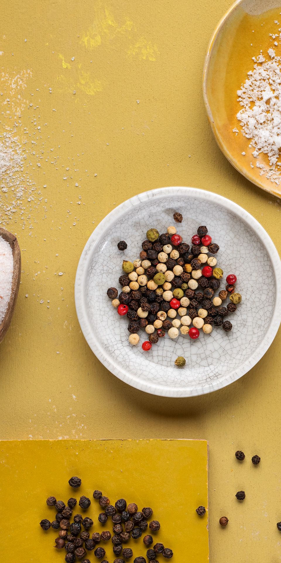 Von oben fotografierte WIBERG Pfeffer- und Salzsorten: Rosa Ursalz, weißes Meersalz und bunte Pfeffersorten locker auf weißen und gelben Tellern arrangiert auf hellem Hintergrund.